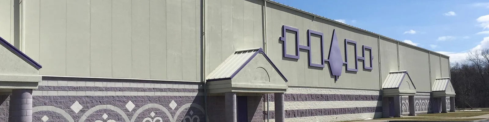 El largo muro gris de la arena con el emblema morado del Hiawatha Belt y dos marquesinas de entrada en punta, visto en una amplia franja desde el estacionamiento. — Onondaga Nation Arena