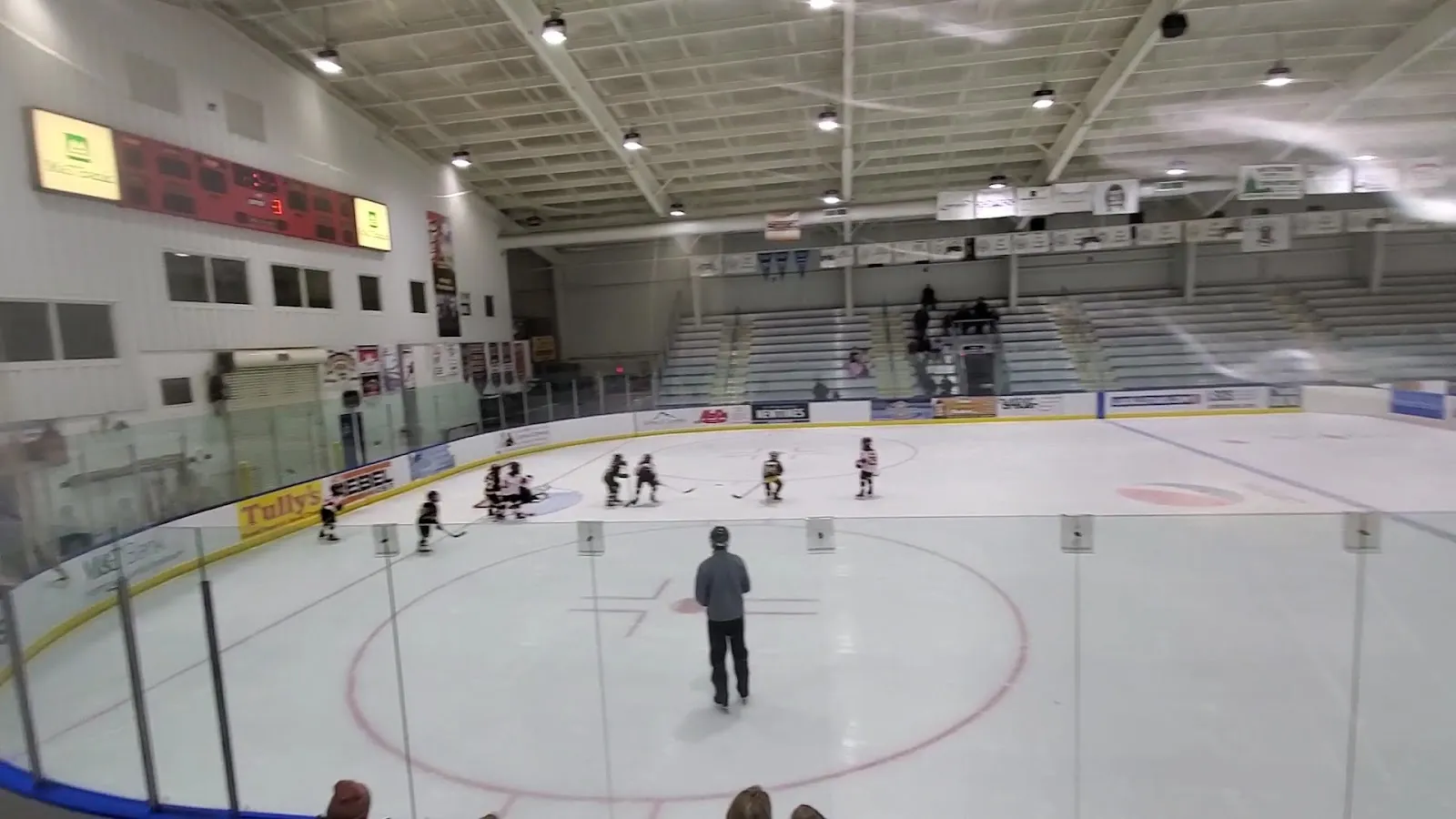 El hielo de Onondaga Nation Arena desde detrás del cristal durante una sesión de hockey juvenil, con asientos y un marcador bajo el techo de acero blanco. — Onondaga Nation Arena