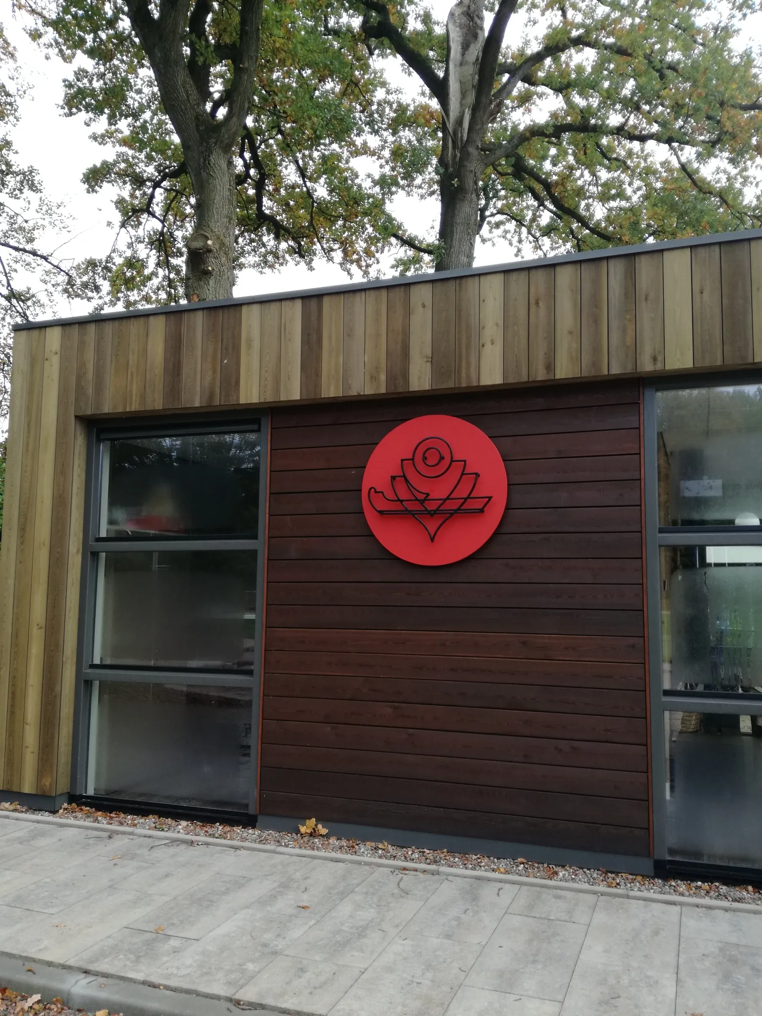 Het met hout beklede clubgebouw van de Oosterbeekse IJsvereniging met het rode embleem van de club op de wand, gezien vanaf het bestrate pad. – Oosterbeekse IJsvereniging