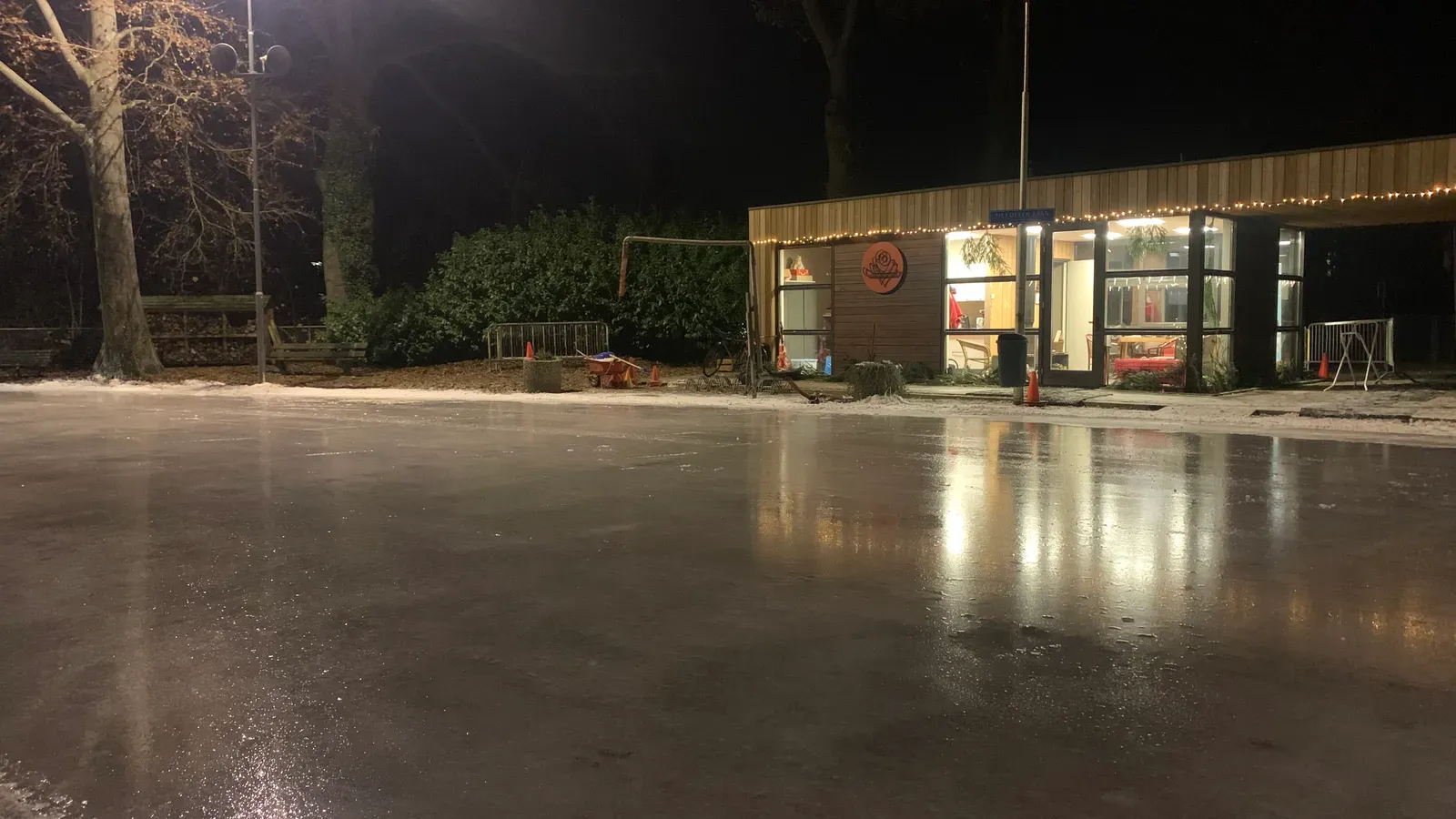 Het gladde, verlichte natuurijs van Oosterbeek 's avonds, met het verlichte clubgebouw en bomen langs de overkant. – Oosterbeekse IJsvereniging