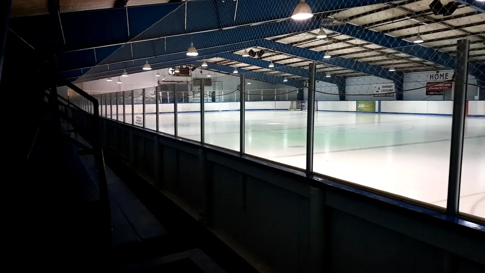 El hielo de Optimist Arena visto desde las gradas a oscuras, mirando a lo largo del vidrio hacia la banca local bajo un techo de acero azul. — Optimist Arena