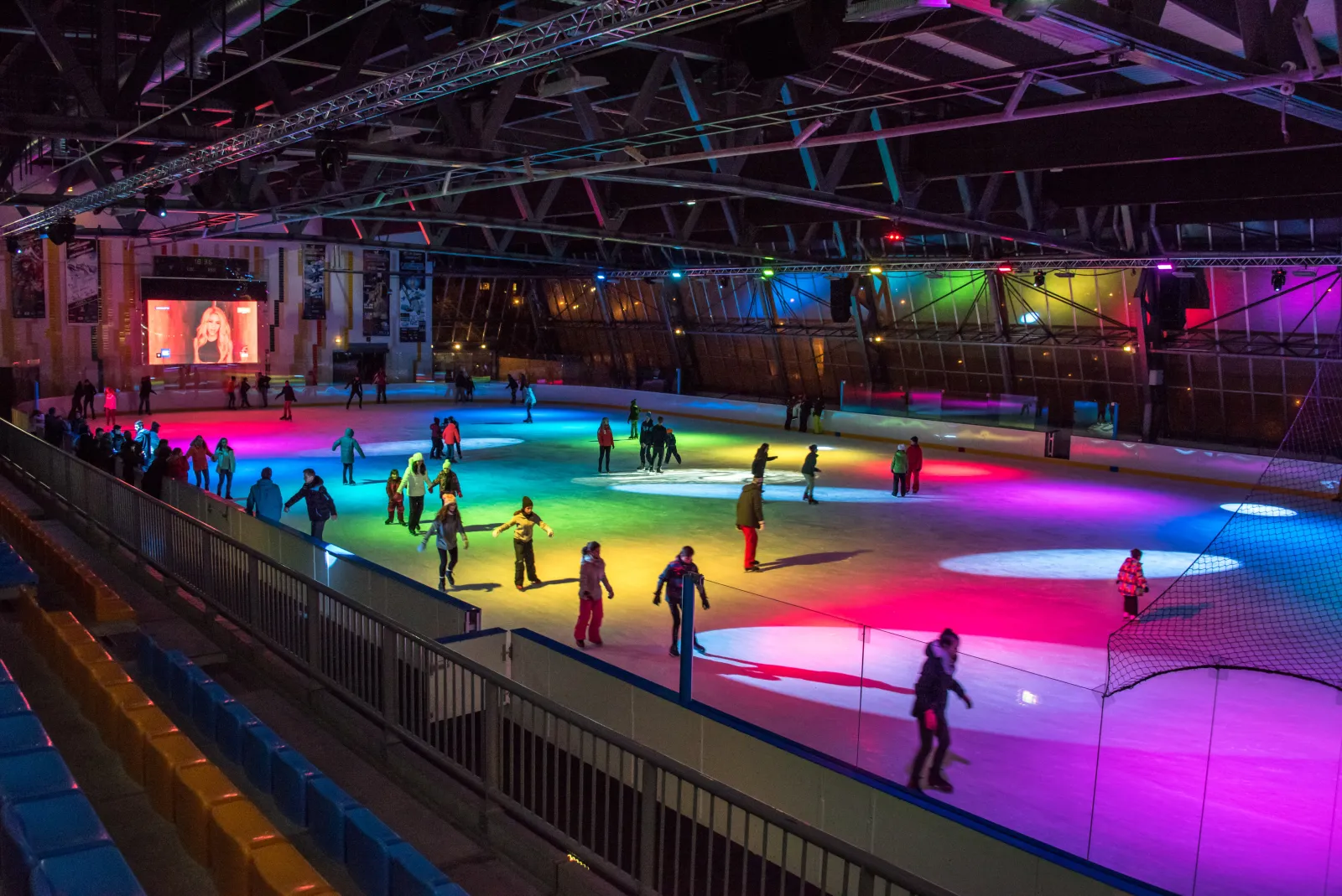 Des patineurs sur la glace éclairée en couleurs de la patinoire de l’Orcières Arena 1850, vue depuis les gradins, avec l’écran géant au fond. — Patinoire d'Orcières Arena 1850