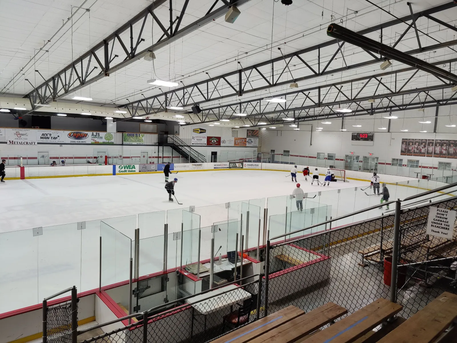 The ice seen from the bleachers behind the glass during a practice, under a white steel-truss roof with banners on the far wall. — Oregon Ice Arena