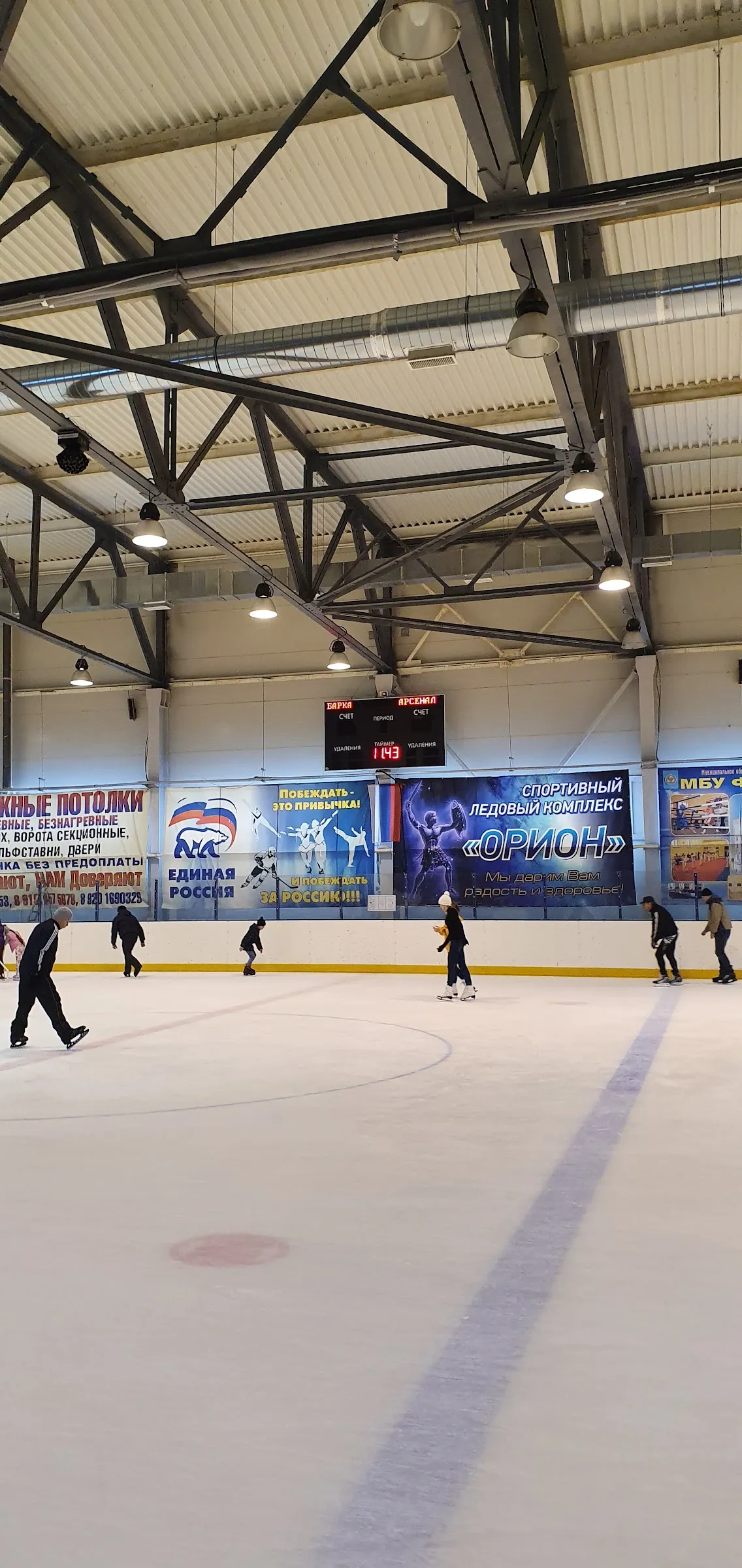 The ice sheet of the Orion sports ice complex in Bologoye seen from the side under steel roof trusses, with a few skaters in the distance — Orion Sports Ice Complex Bologoye