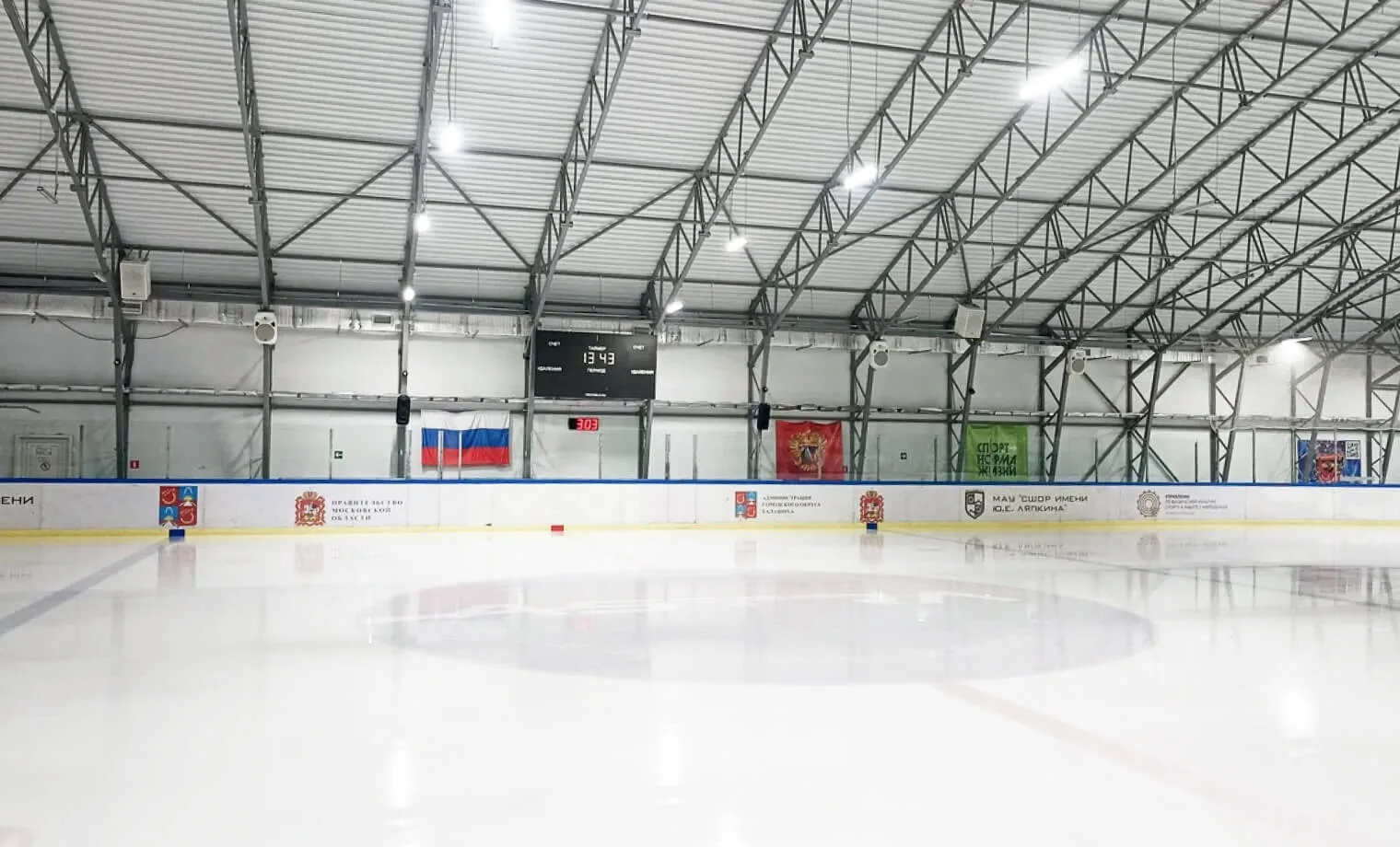 The empty ice sheet of the Orion Technopool Arena in Balashikha under the steel roof trusses, with the boards and the flags on the far wall. — Orion Technopool Arena