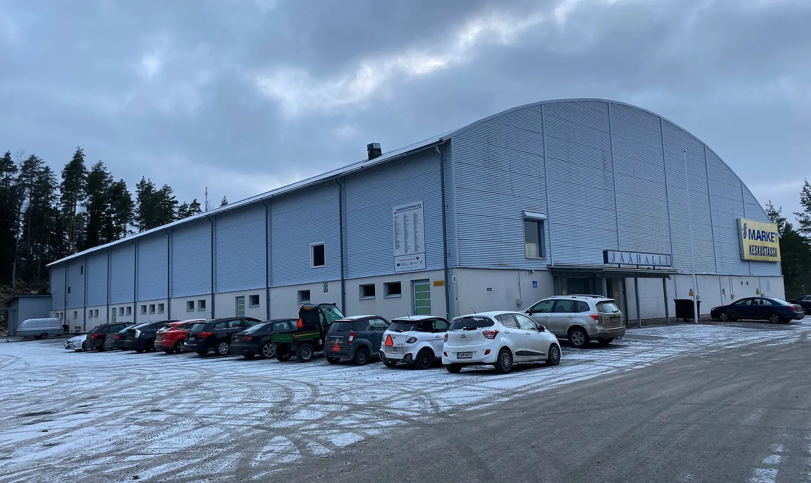 The side and entrance of Oriveden jäähalli, a grey steel-clad hall with a curved roof, seen across the car park under a cloudy winter sky. — Oriveden jäähalli