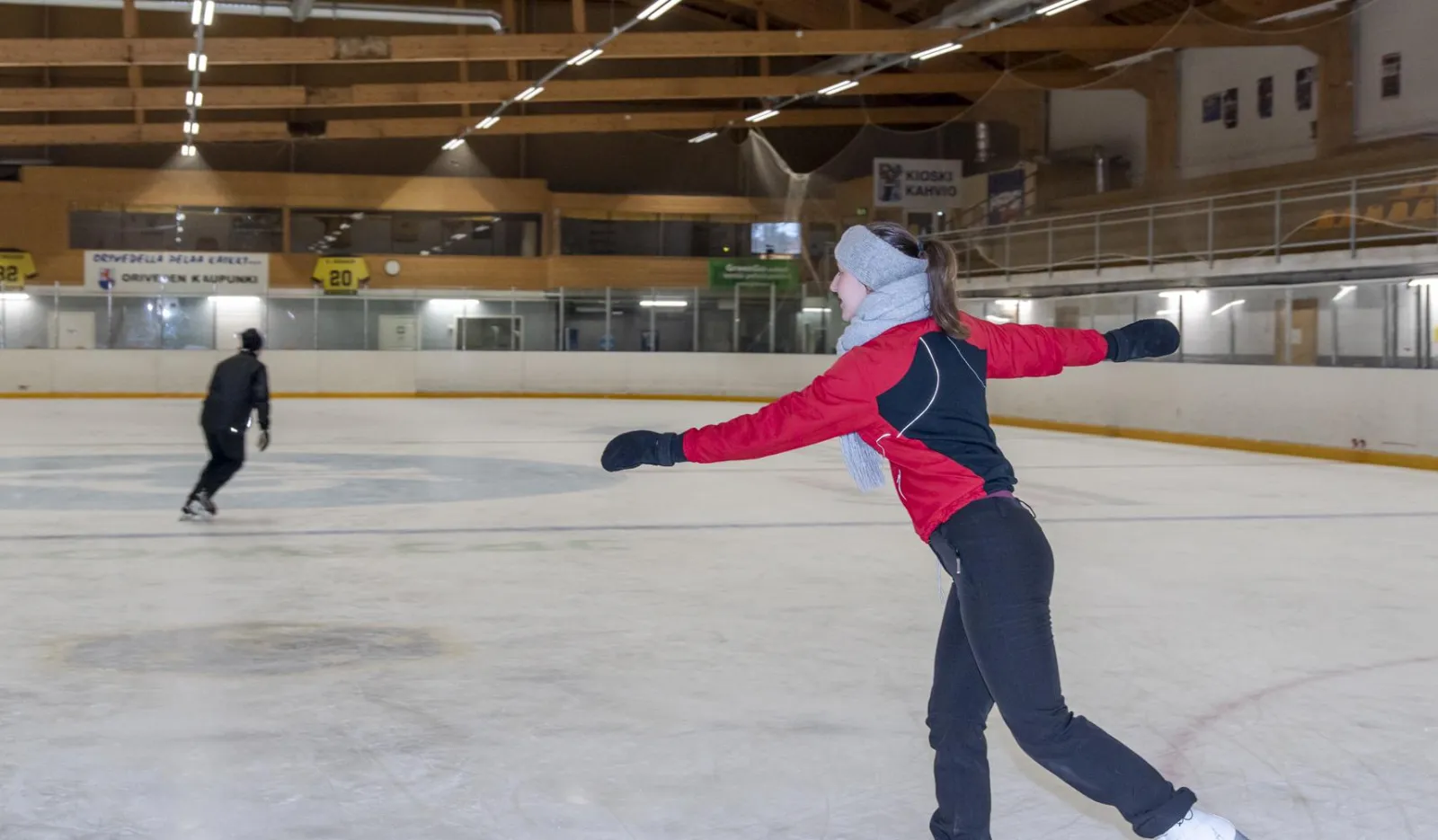Two skaters on the ice of Oriveden jäähalli under a timber roof, one in the foreground with arms outstretched mid-glide seen from behind and one further back. — Oriveden jäähalli