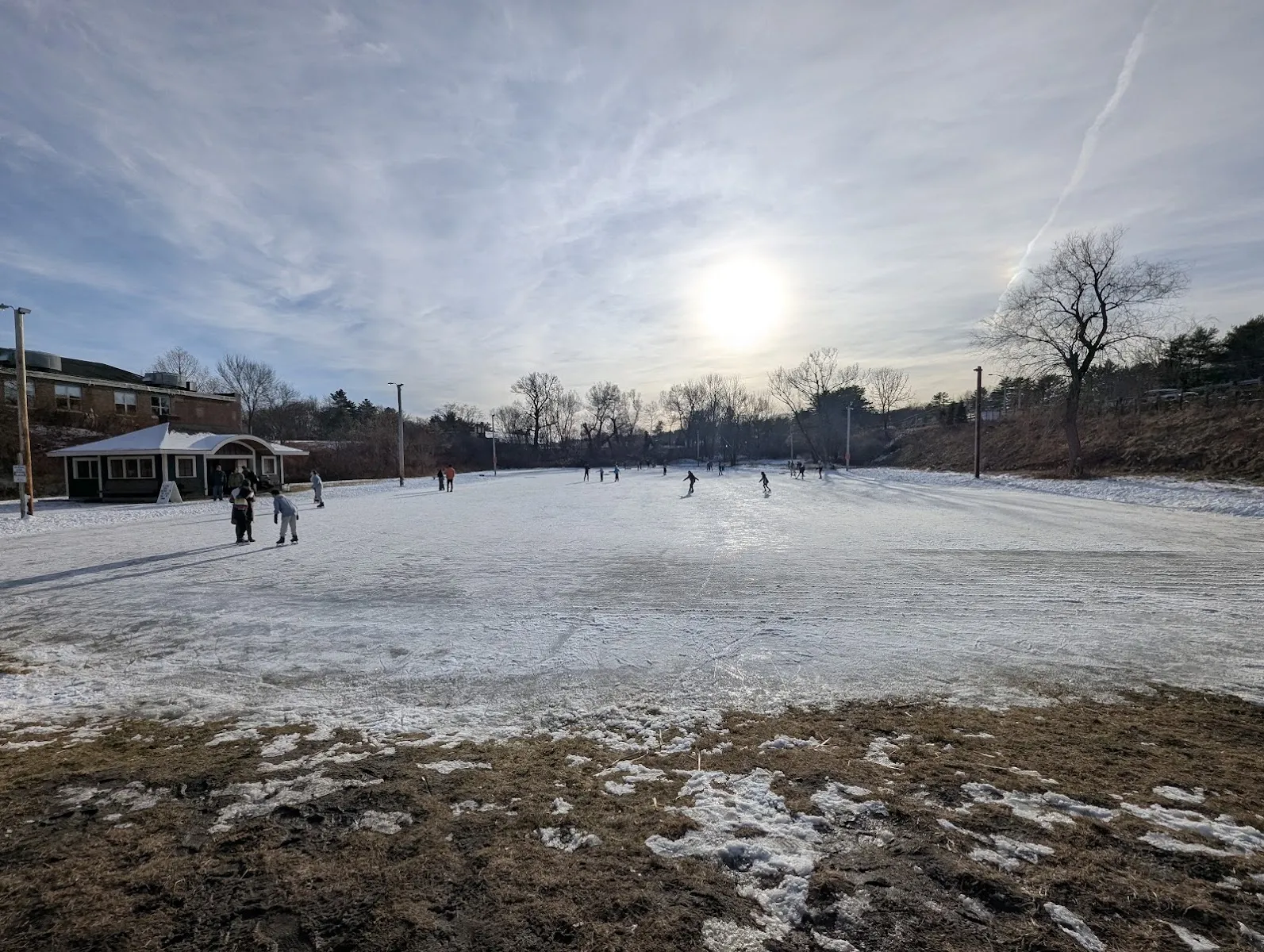 Distant skaters on the frozen Blake Skating Pond with the warming house at the left, under a low winter sun — Orland H. Blake Skating Pond