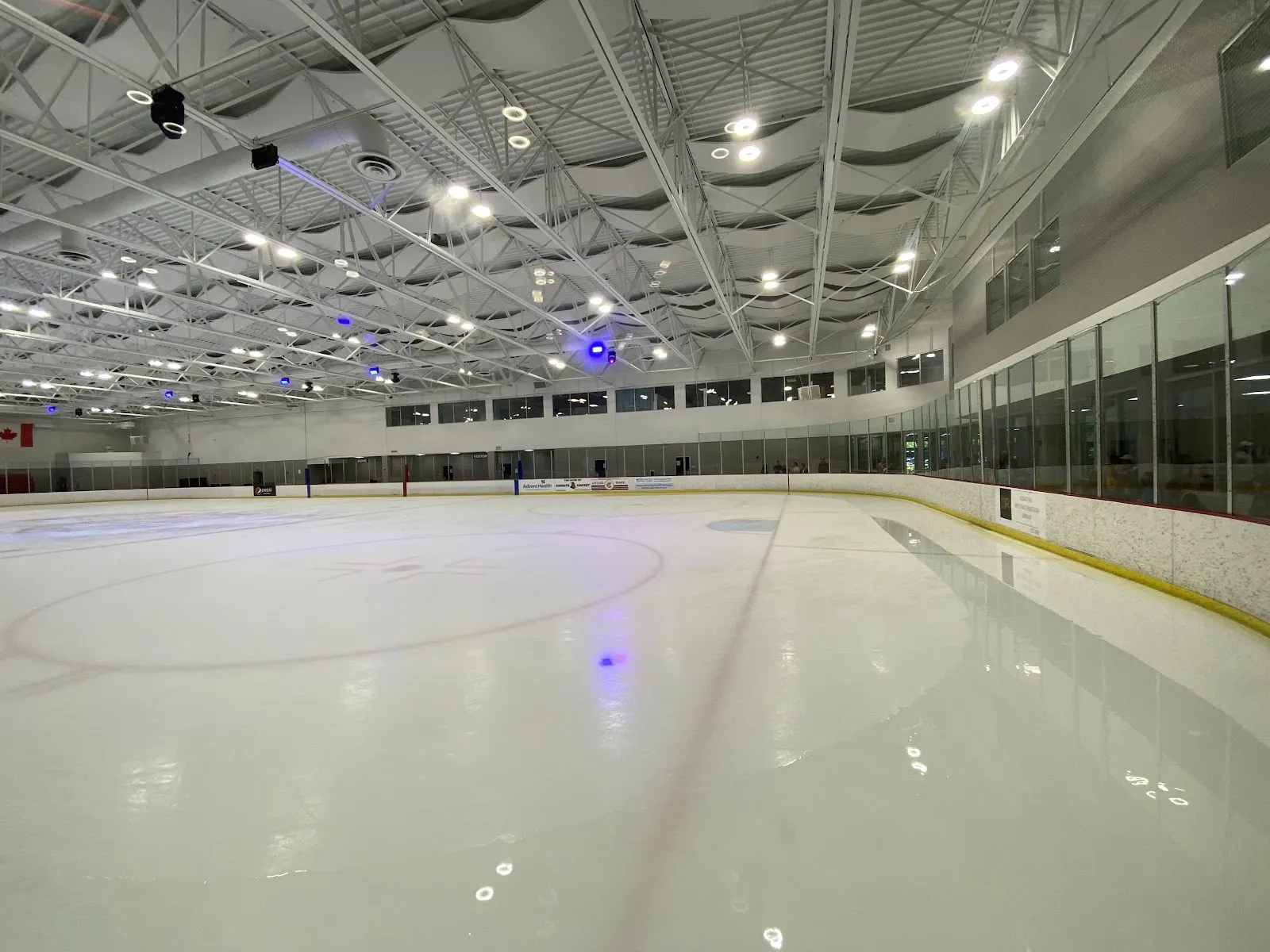 Una pista amplia y vacía vista desde el extremo de las vallas, con hielo blanco, un tenue círculo central, bajo un techo de luces y vidrio verde a lo largo del lado opuesto. — Orlando Ice Den