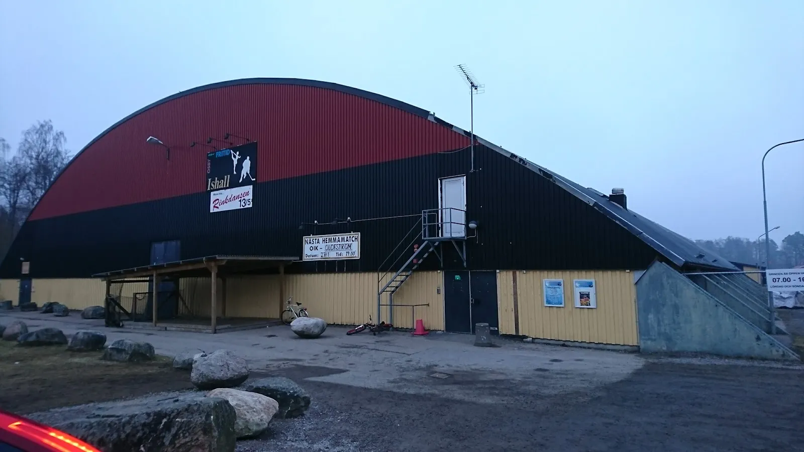 The arched red-and-black roof of Osby ishall with its Ishall sign, seen from the car park — Osby ishall