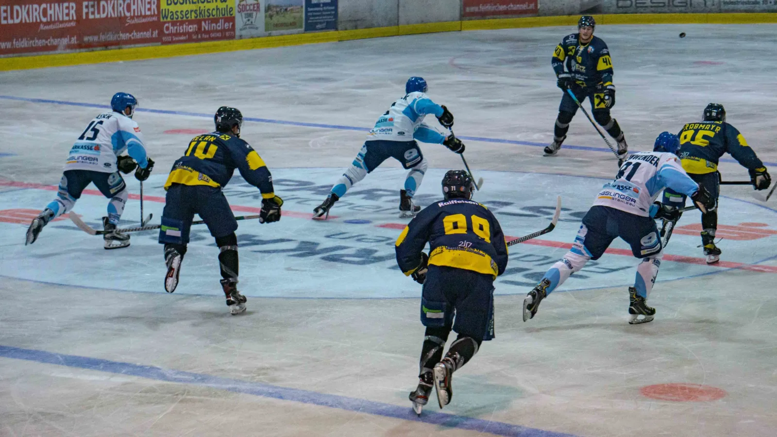 Players in dark blue and yellow and in white and light blue skate across the ice during a game, with advertising boards along the far side. — Ossiacher See Halle