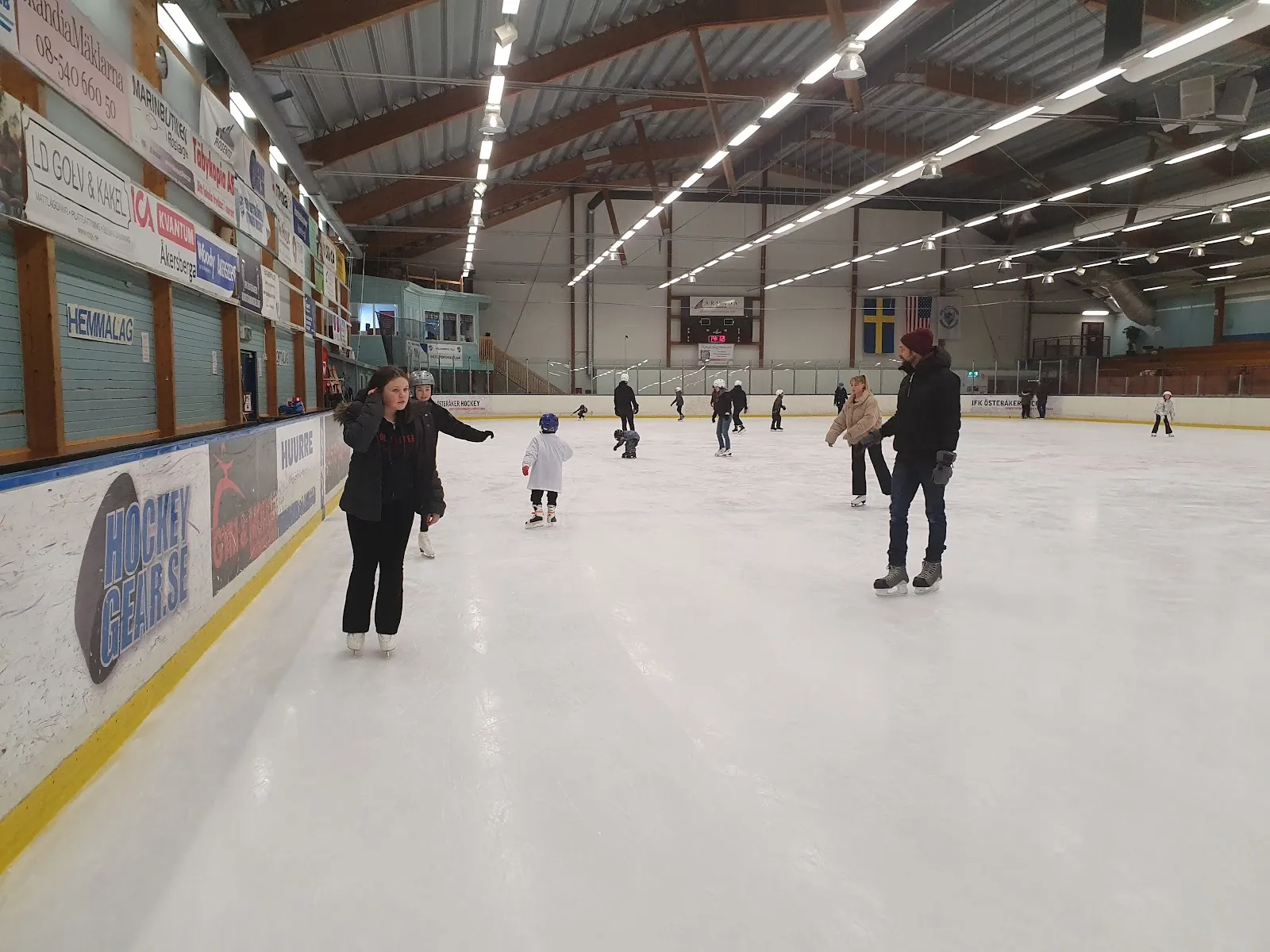 Public skating on the ice sheet in the hall, with skaters of several ages spread along the near boards, advertising panels on the boards and a long steel-framed roof with rows of lights overhead. — Österåkers ishallar