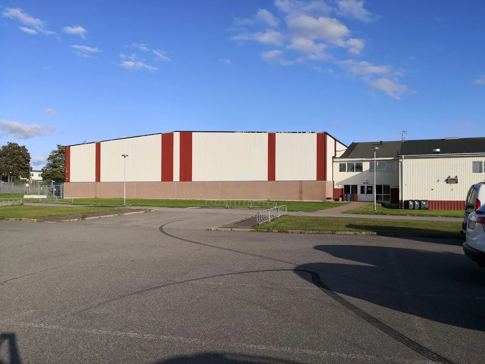 The red-striped steel hall of Österås ishall in Hässleholm seen across the car park under a blue sky — Österås ishall