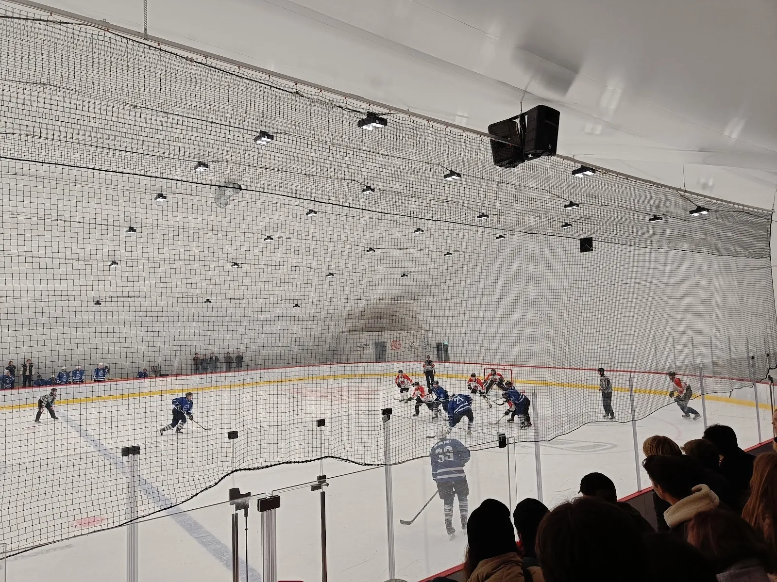 En match på isen i Östermalms ishall i Stockholm, sedd från läktaren över isytan under halltaket, med åskådare längs sargen. – Östermalms ishall