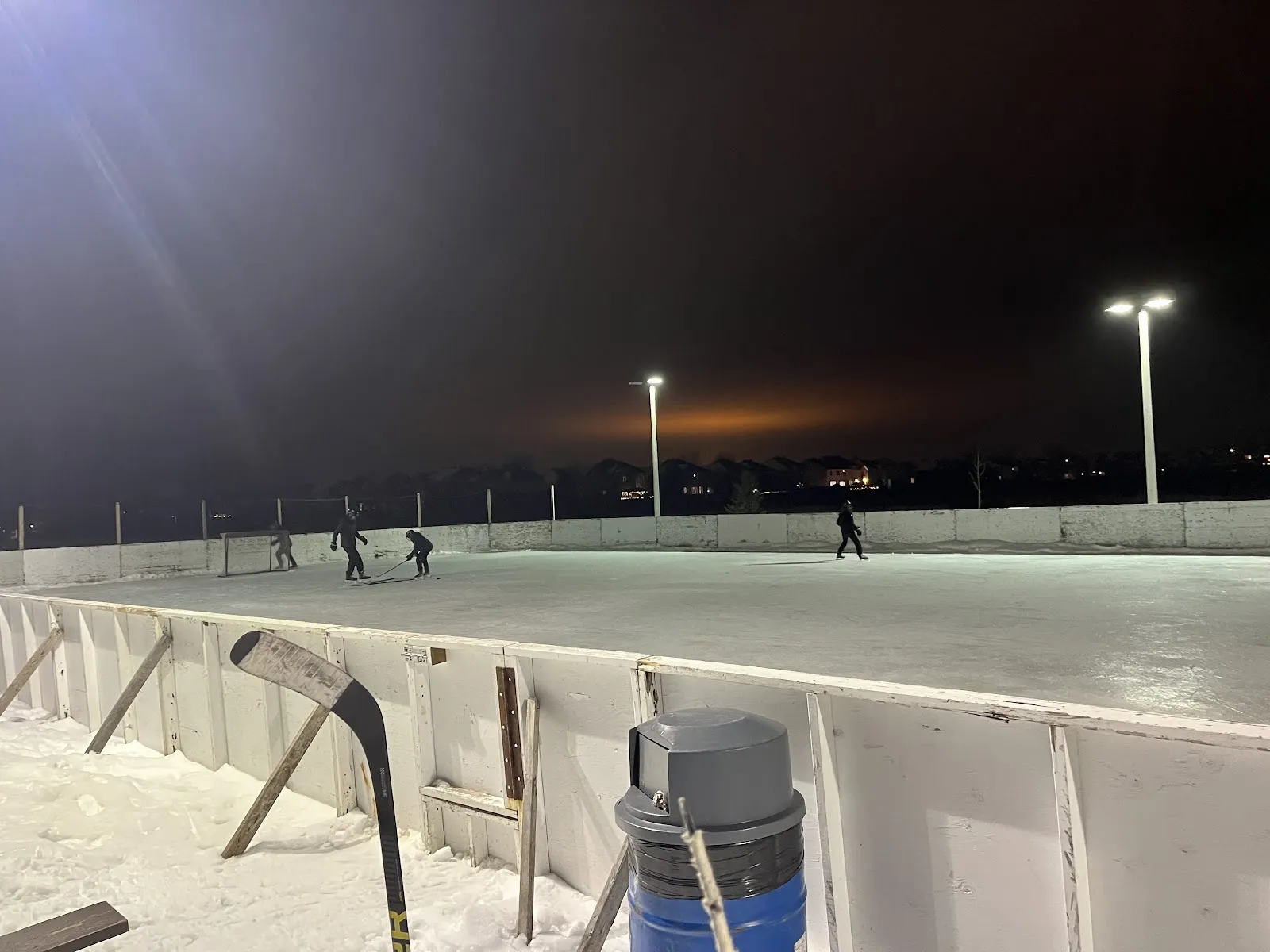 The Aquaview Park rink at night, a few players spread across the ice beside a hockey net with white boards, floodlights and a stick leaning on the rail. — Aquaview Park Outdoor Rink