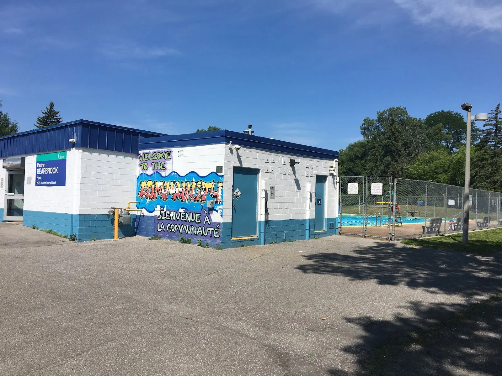 El edificio blanco y azul de la alberca de Bearbrook Park con su letrero y su mural de bienvenida, visto al otro lado del estacionamiento en un día soleado, con la cerca de la alberca a la derecha — Bearbrook Park and Pool Outdoor Rink