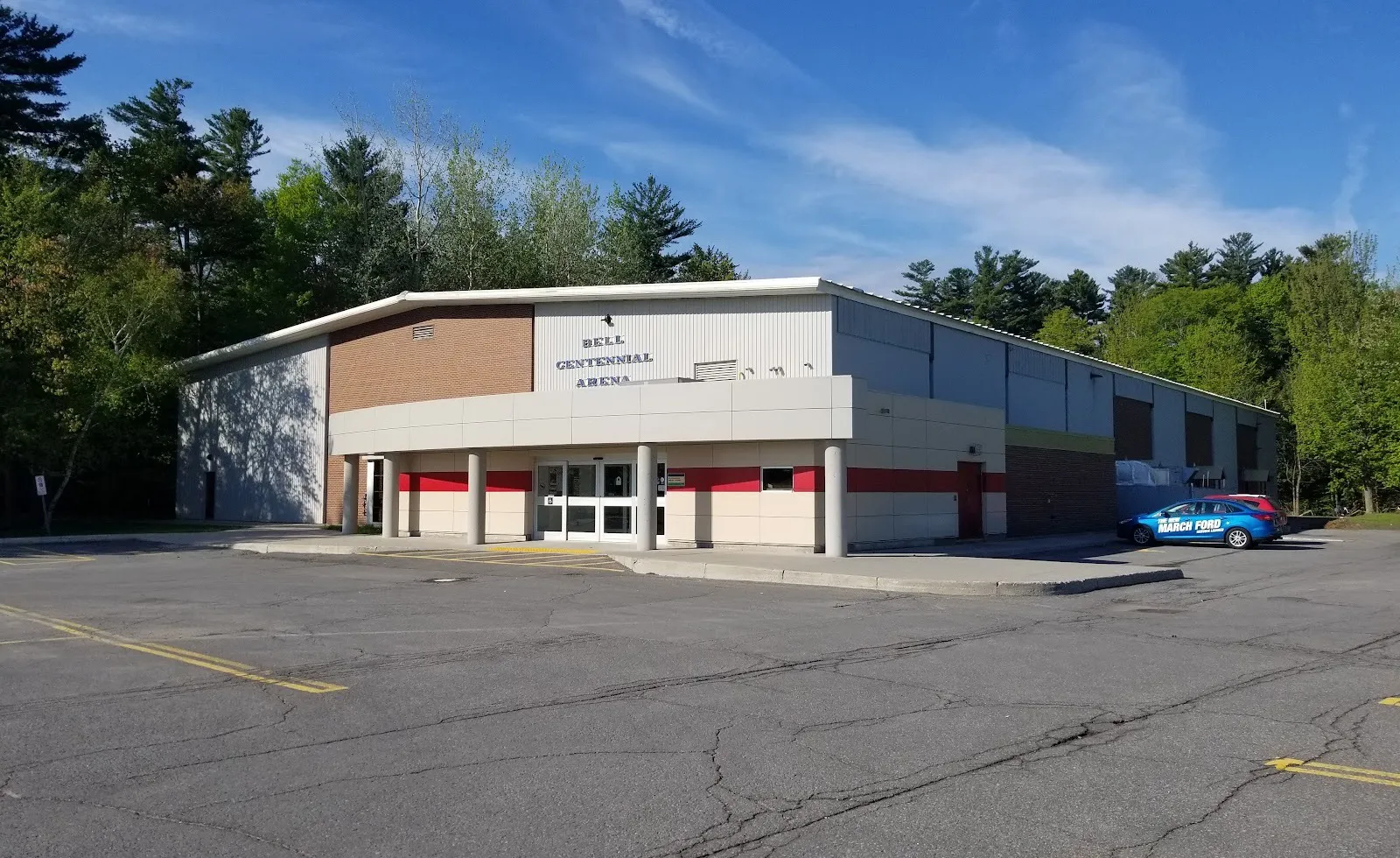 La Bell Centennial Arena en Cassidy Road, en Ottawa, con su nombre en letras sobre la entrada con columnas y pinos detrás — Bell Centennial Arena