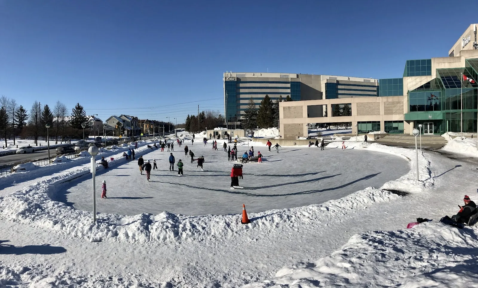 Skaters spread across the outdoor rink at Ben Franklin Place beneath a blue sky, the civic centre's glass front and snow banks behind the ice. — Ben Franklin Place Outdoor Rink