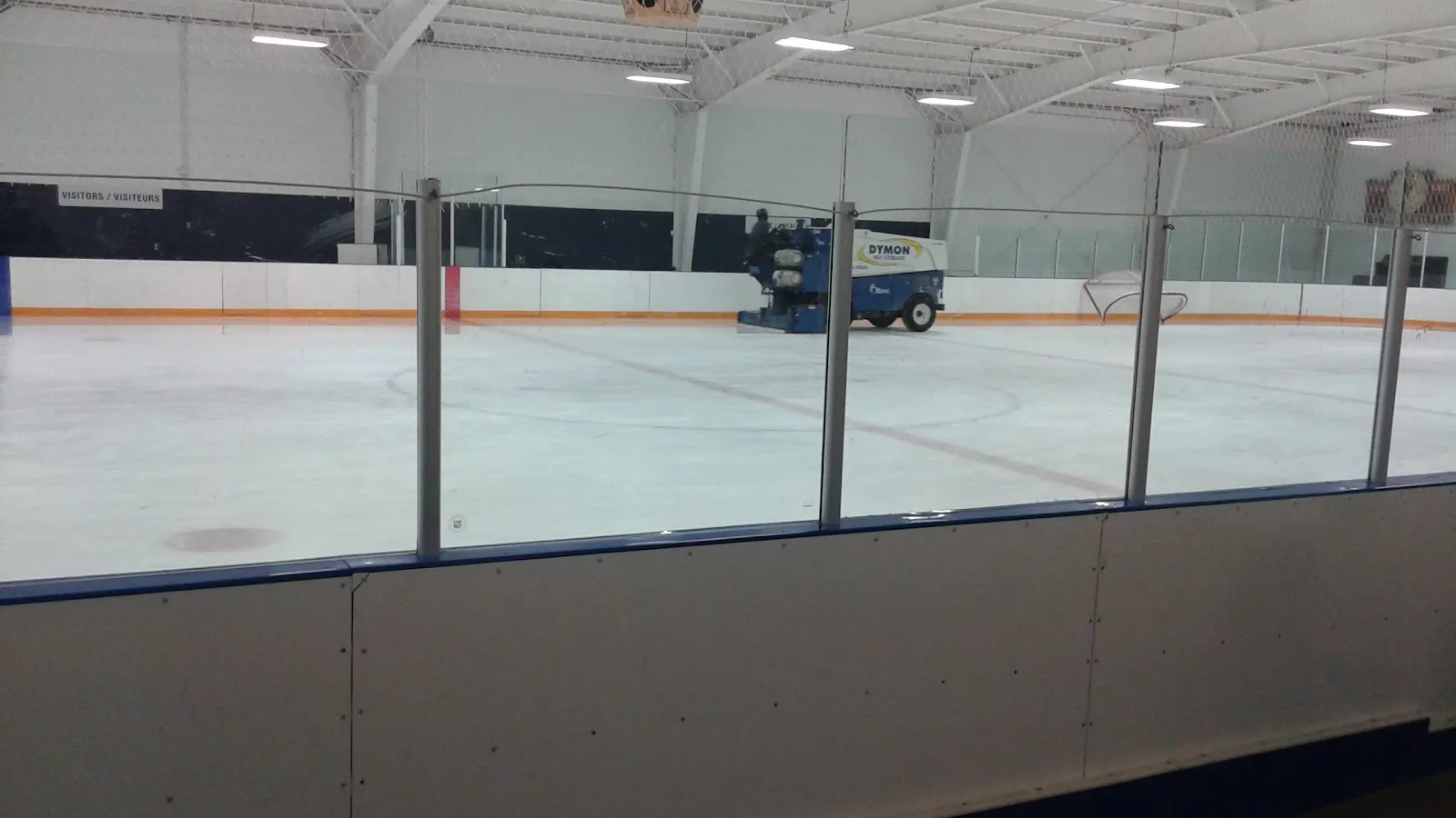 El hielo vacío de Bernard Grandmaître Arena visto sobre las vallas y el cristal, con la pulidora estacionada contra las vallas del fondo bajo el techo blanco de acero — Bernard Grandmaître Arena