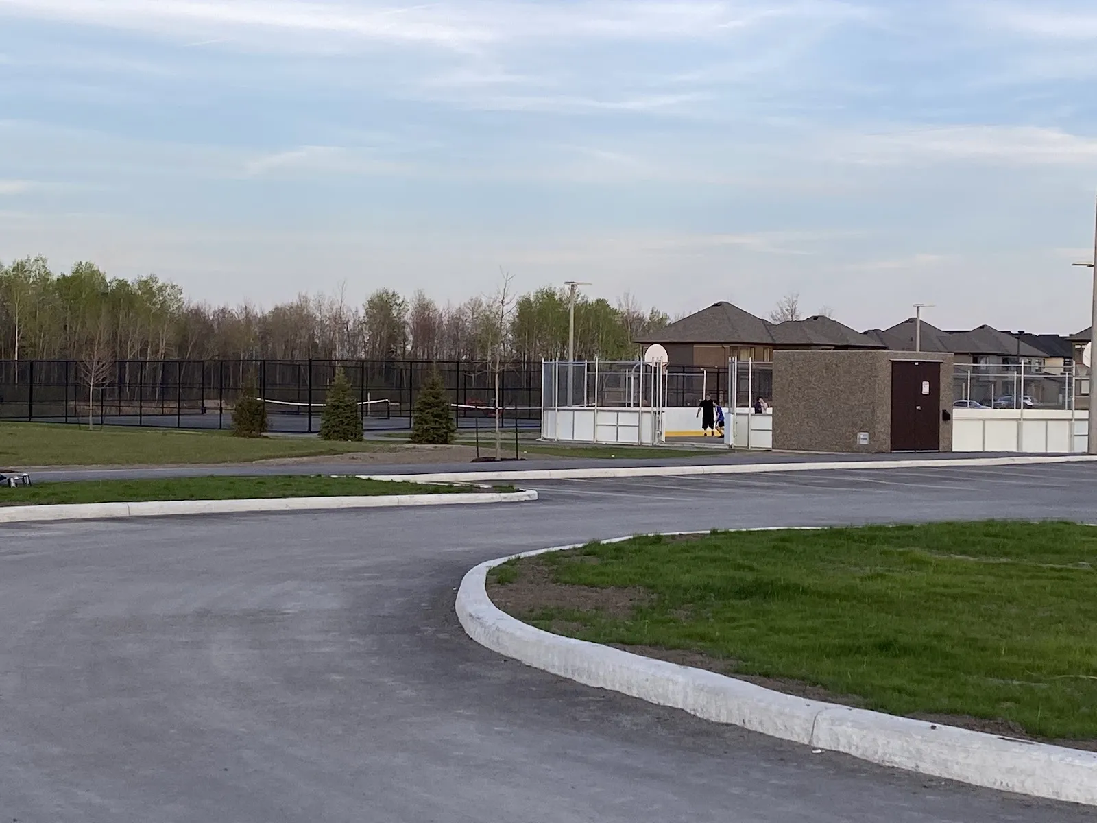 Bert Dowler Park's boarded court and small service building beyond a paved loop, with tennis courts behind. — Bert Dowler Park Outdoor Rink