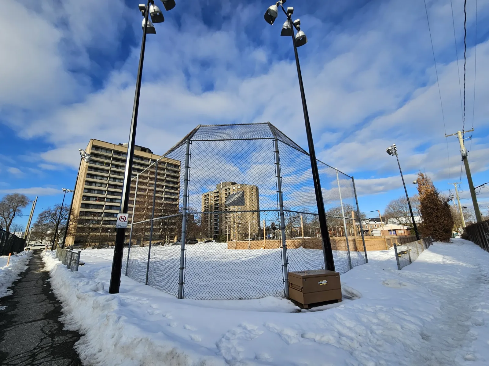 Bingham Park in winter: a snow-covered fenced court under tall light standards, with apartment towers and houses around the park. — Bingham Park Outdoor Rink