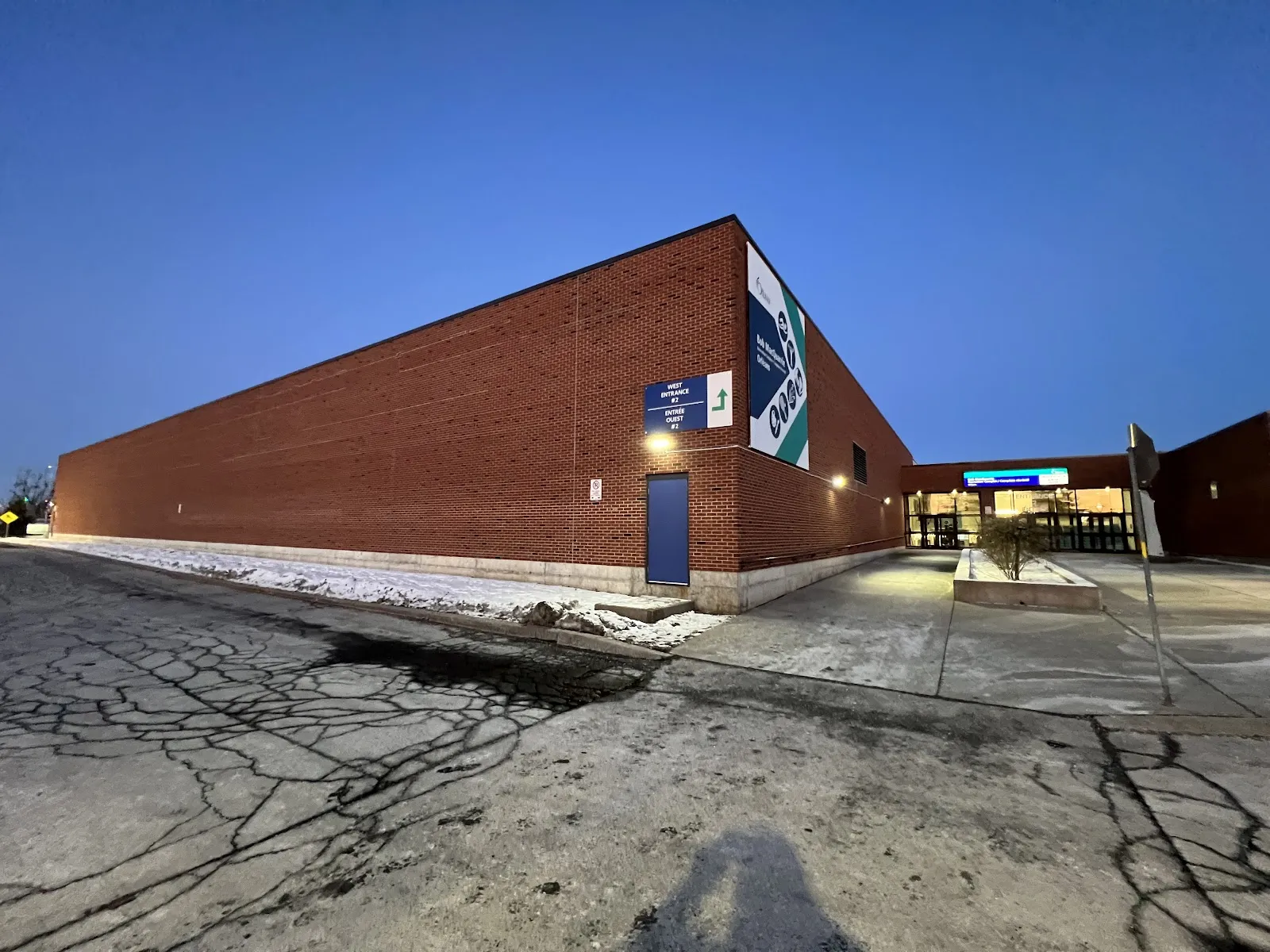 El Bob MacQuarrie Recreation Complex al atardecer, un largo muro de ladrillo rojo con una manta grande y, más allá, la entrada de cristal iluminada. — Bob MacQuarrie Recreation Complex – Orléans
