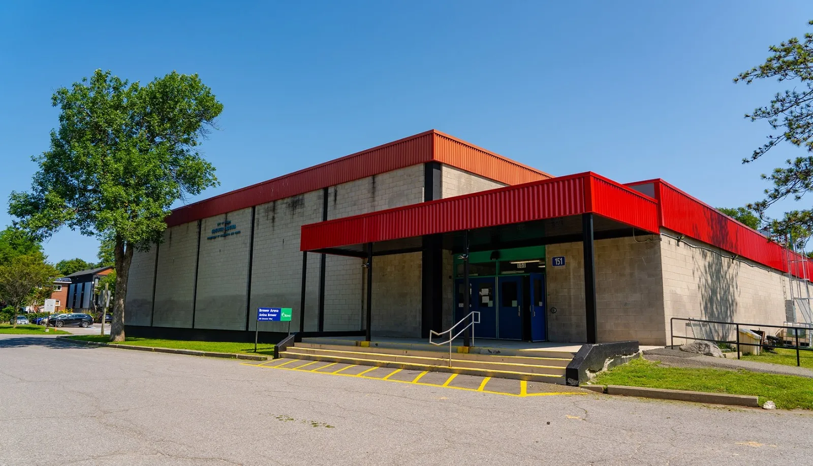 Entrada principal de Brewer Arena, un edificio de bloques beige con una marquesina metálica roja sobre las puertas y un letrero verde de 'Brewer Arena / Aréna Brewer' junto a la rampa accesible. — Brewer Pool and Arena
