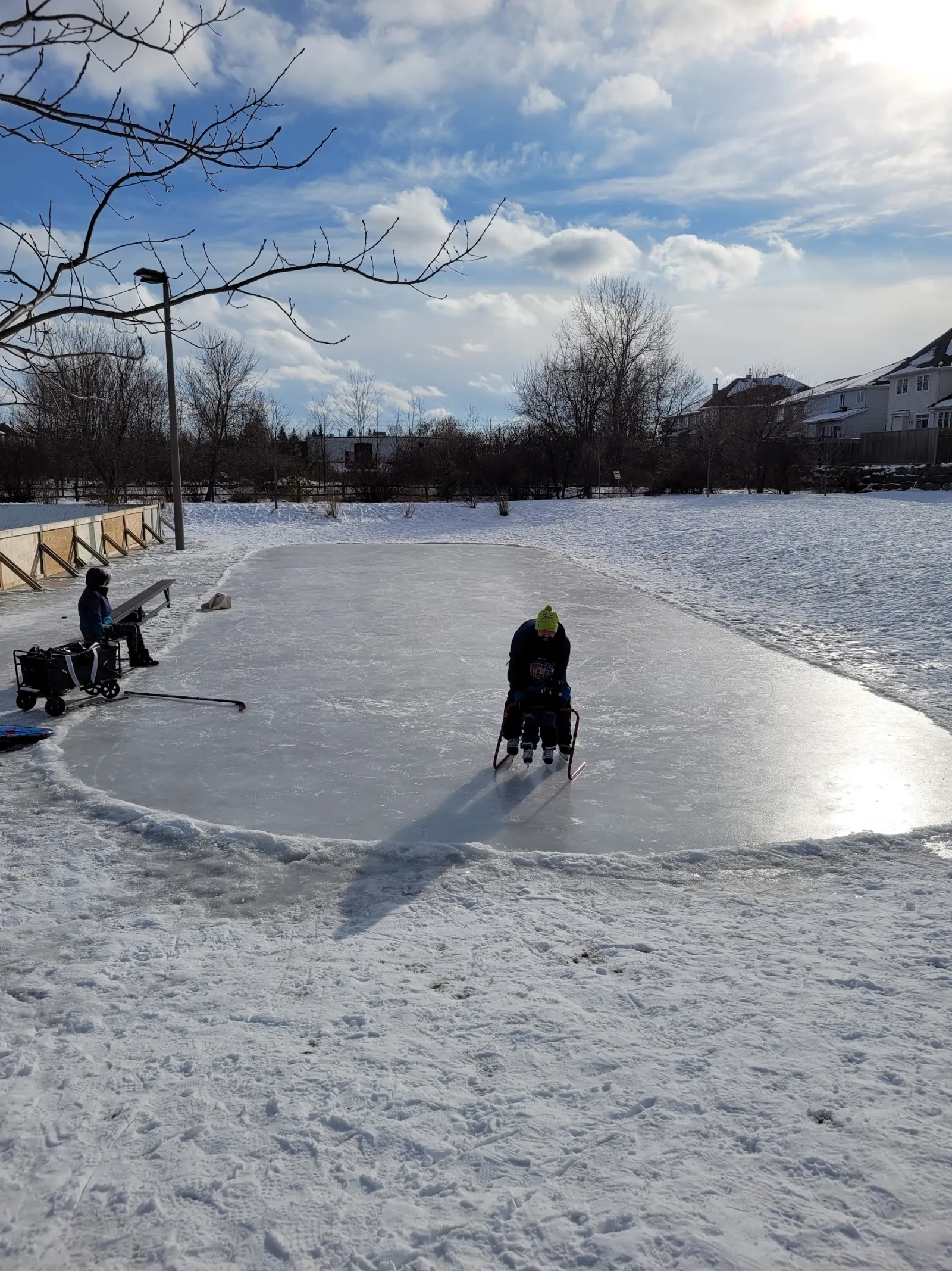 The park's outdoor rink in winter: a flooded oval of ice inside low boards, with a couple of skaters and snowbanks around it under a bright sky. — Bryanston Gate Park Outdoor Rink