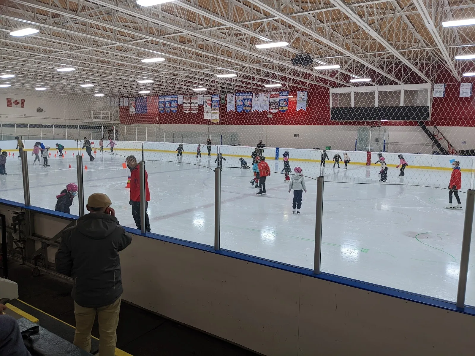 Una sesión pública vista a través del vidrio, con patinadores repartidos por la pista bajo un techo de armaduras de acero con banderines rojos en la pared del fondo. — Canterbury Recreation Complex – Brian Kilrea Arena