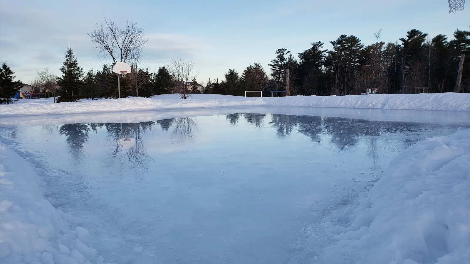 Una pista inundada al aire libre al atardecer, una amplia superficie lisa que refleja el cielo y los árboles sin hojas, con bancos de nieve a lo largo de los bordes y una canasta de baloncesto al fondo. — Cardinal Creek Community Park Outdoor Rink