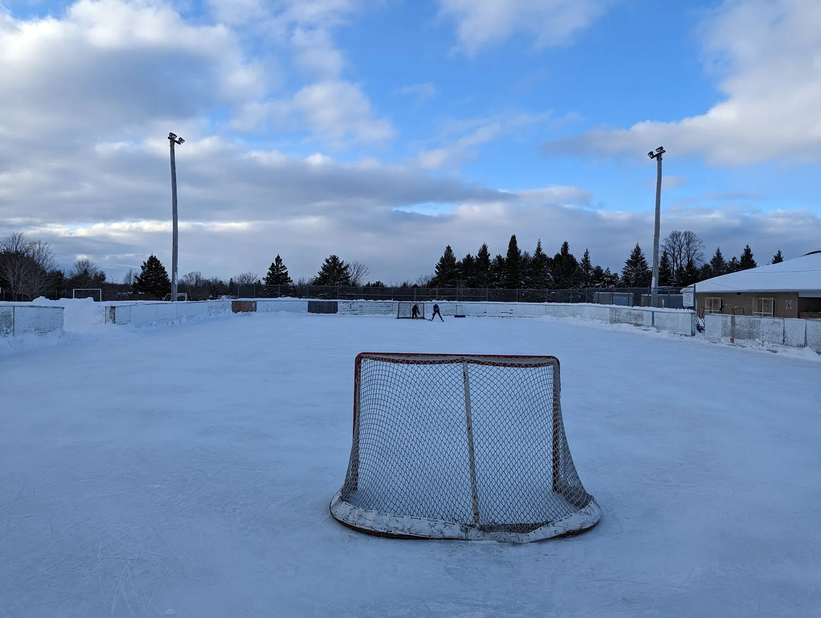The Corkery community park rink under a blue sky, a hockey net on the snowy flooded surface with light poles and a low building at the far side. — Carrol Homestead Community Park / Corkery Community Centre Outdoor Rink