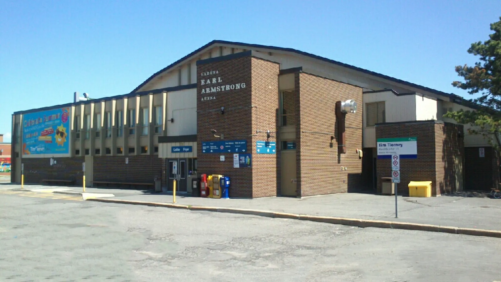 Brick and beige-panel arena building from the parking lot, with bilingual signage reading "ARENA EARL ARMSTRONG ARENA" over the main entrance and a lobby/foyer sign. — Earl Armstrong Arena