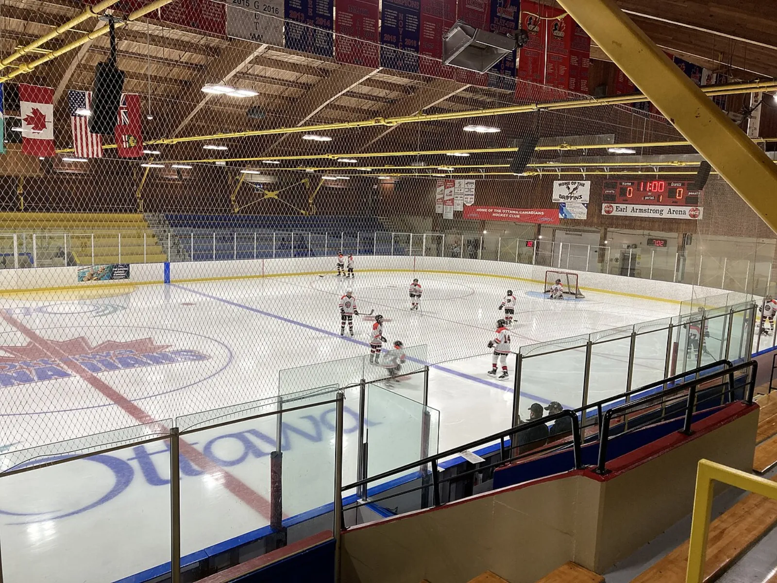 Wide view of the full ice sheet from the upper stands, with an "Earl Armstrong Arena" scoreboard sign, Canada/USA/Ontario flags, championship banners, and a youth hockey game underway on the ice. — Earl Armstrong Arena