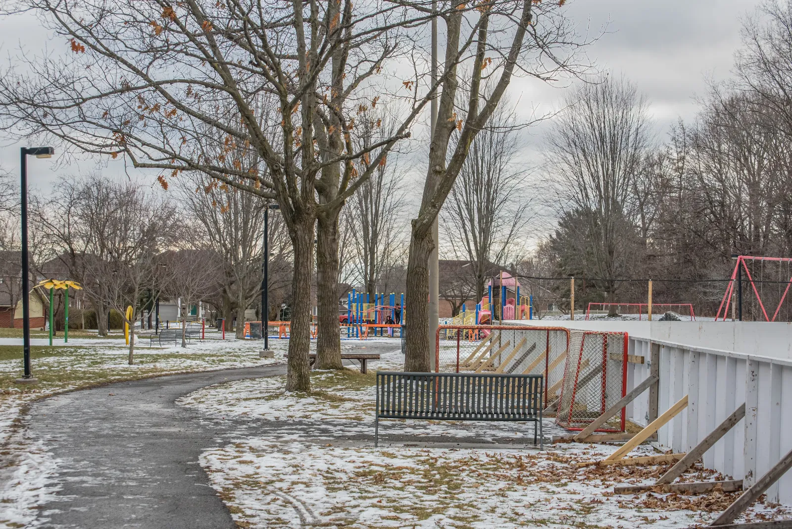 Fairfield Park a finales del otoño con una ligera capa de nieve: las vallas blancas de la pista al aire libre junto a un sendero y una banca, con el área de juegos detrás. — Fairfield Park Outdoor Rink