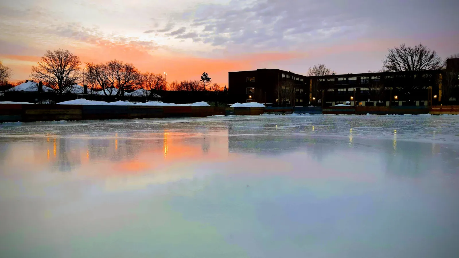 The Fisher Park outdoor rink at sunset, its freshly flooded ice reflecting an orange sky, with the school building and bare trees beyond the boards. — Fisher Park Outdoor Rink