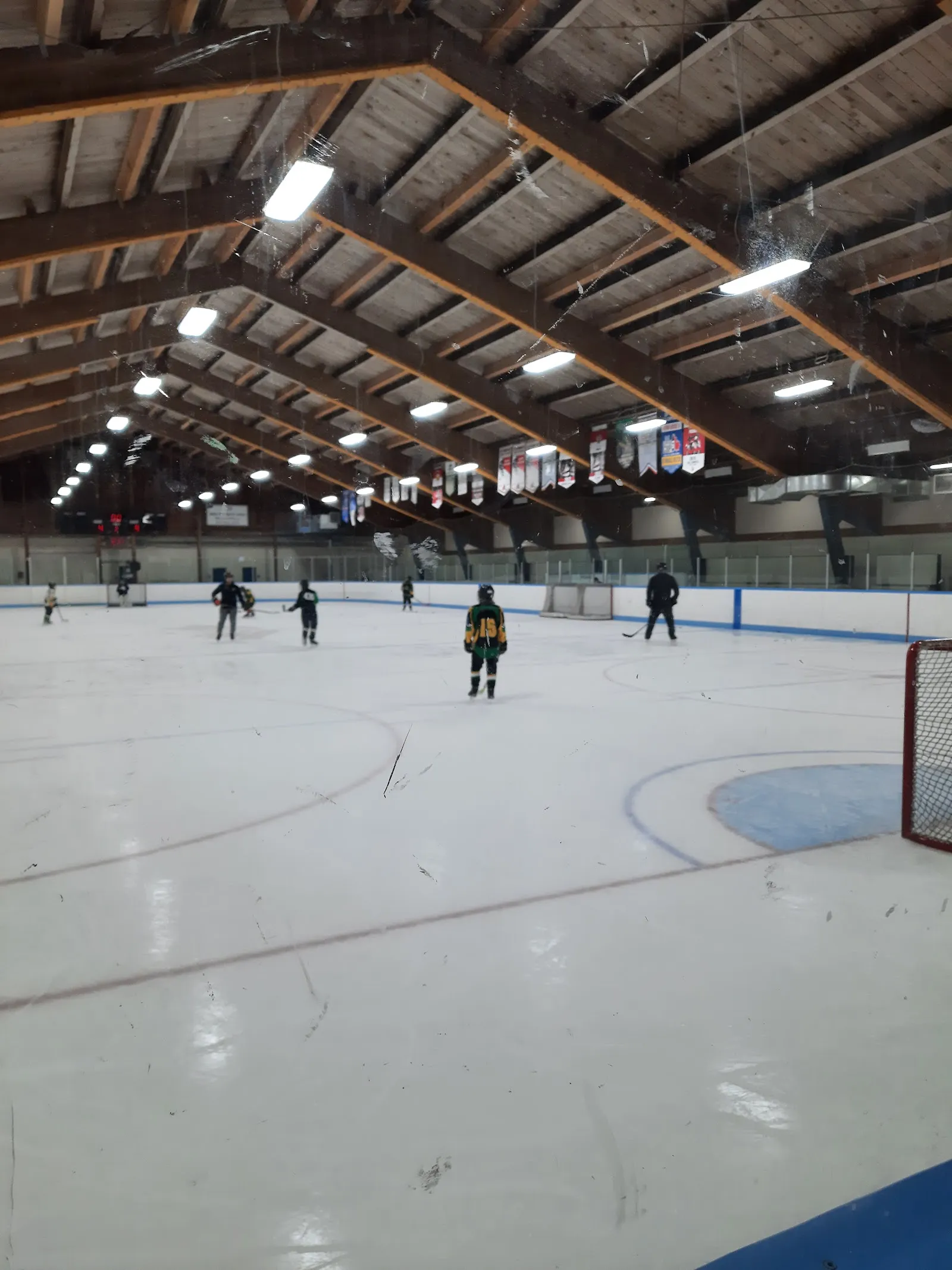 Interior de la pista visto desde las vallas: techo de armaduras de madera, mantas de equipos colgadas de las vigas y varios jugadores en el hielo cerca de una portería. — Fred Barrett Arena
