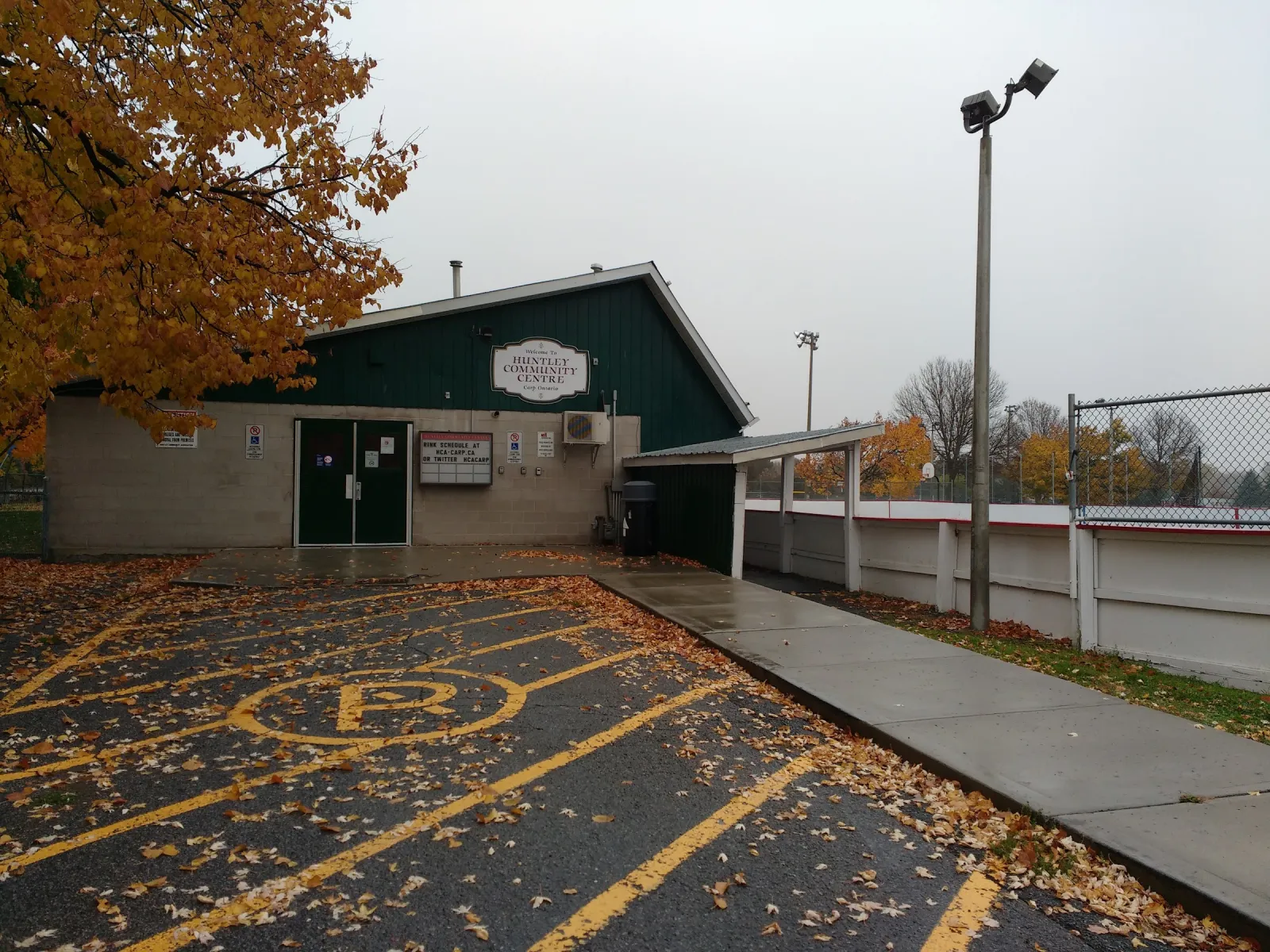 The green-and-white Huntley Community Centre in autumn, with the white boards of the outdoor rink visible behind the fence to the right. — Huntley Community Centre Outdoor Rink