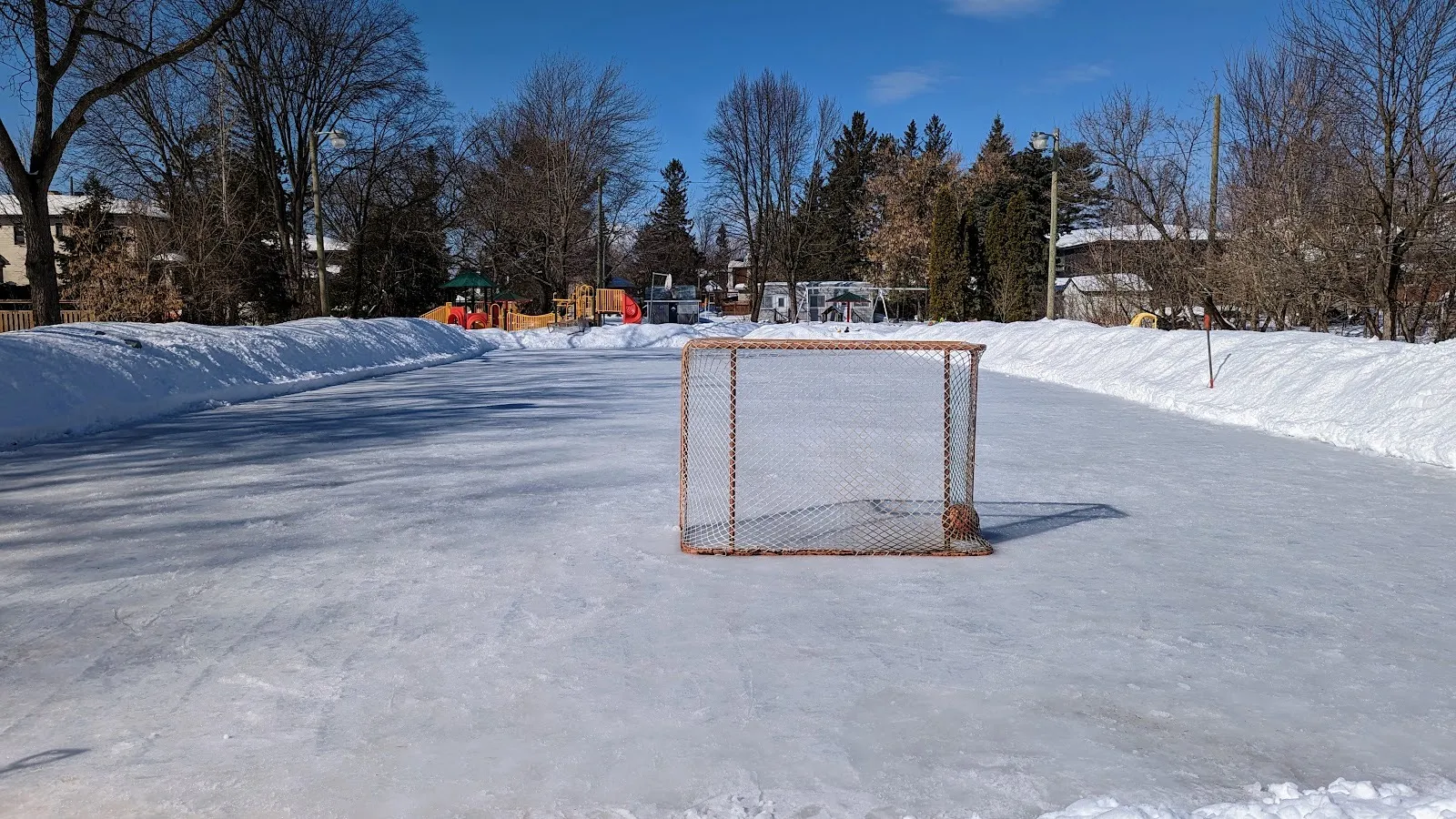 La pista al aire libre inundada de Iona Park, en Ottawa, en un día soleado de invierno, con una portería de hockey en el hielo y bancos de nieve a los lados — Iona Park Outdoor Rink