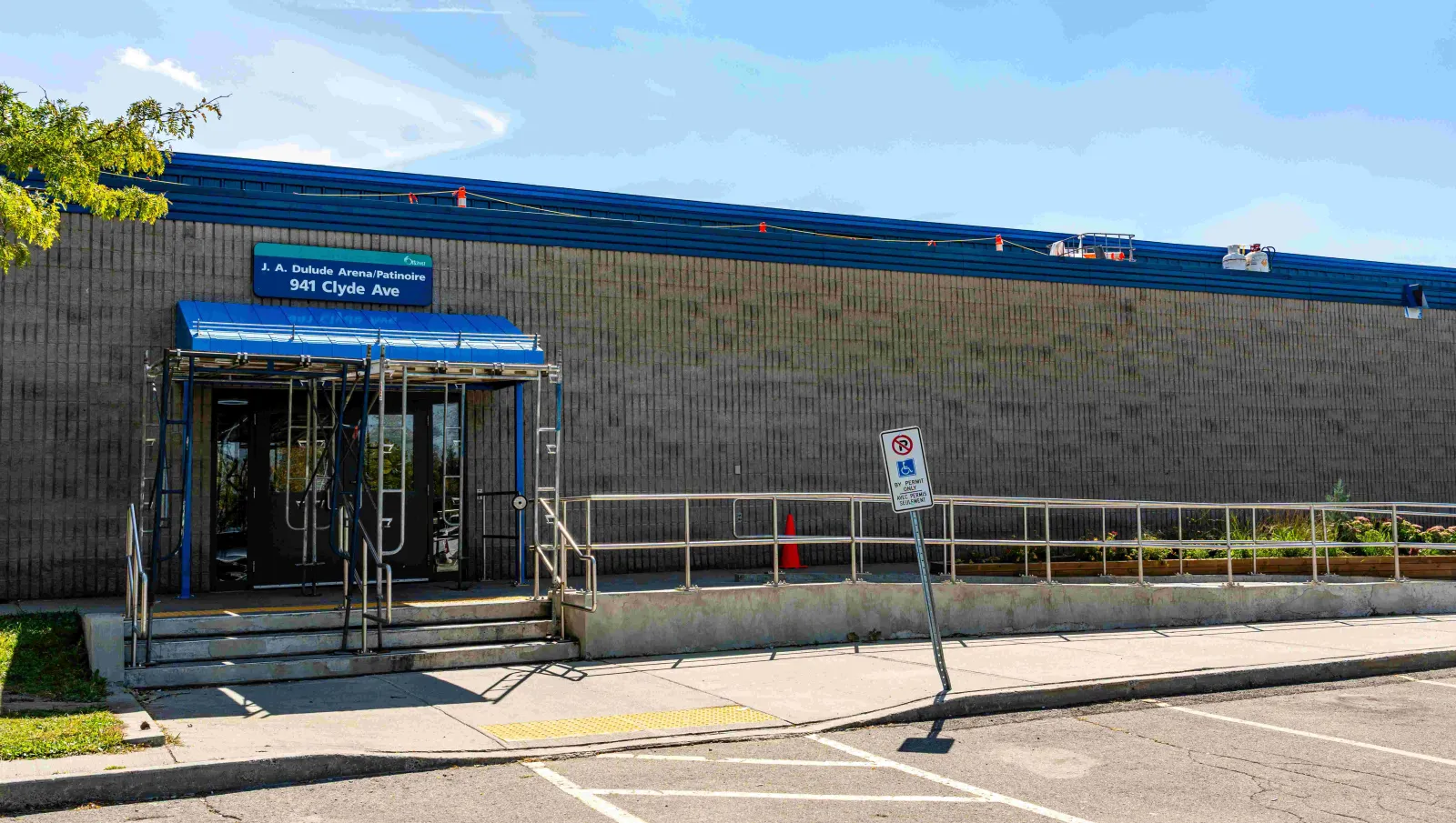 Entrada de J.A. Dulude Arena, un edificio de bloques con remate azul y un letrero azul de 'J. A. Dulude Arena/Patinoire, 941 Clyde Ave' sobre una marquesina y una rampa accesible. — J.A. Dulude Arena