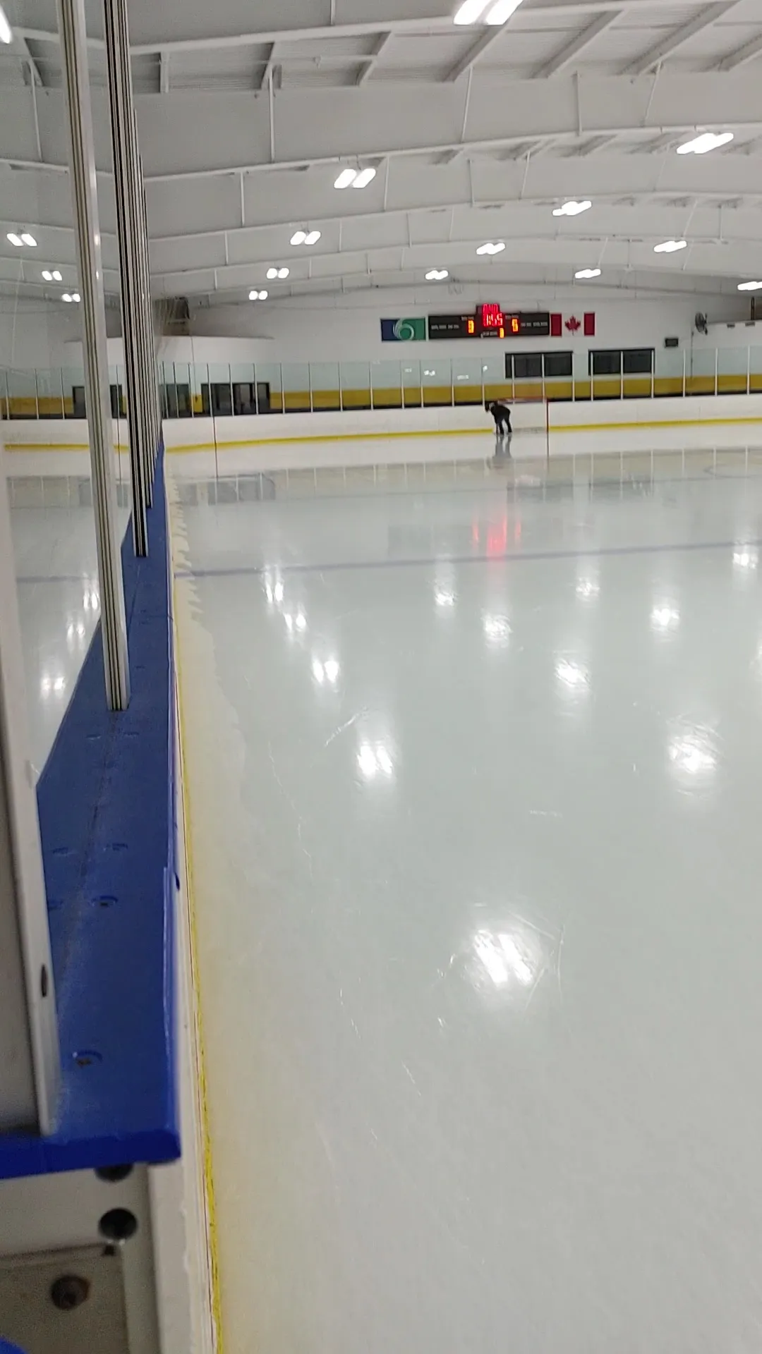Una vista estrecha a lo largo de las vallas de una pista luminosa, con un barandal azul a la izquierda y un marcador al fondo del hielo. — J.A. Dulude Arena