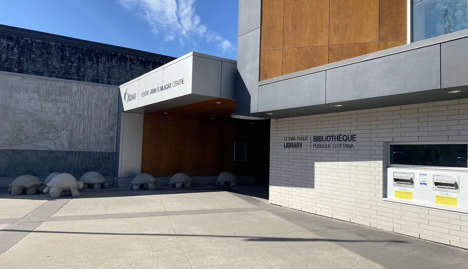 La entrada del John G. Mlacak Centre en Kanata, bajo su marquesina con el nombre junto al ala de la biblioteca, con tortugas de piedra a lo largo del muro — John G. Mlacak Centre