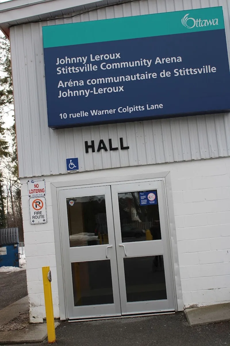 The Hall entrance of Johnny Leroux Stittsville Community Arena: a pair of glass doors in the white wall under the City of Ottawa sign naming the arena in English and French with its address, 10 Warner Colpitts Lane. — Johnny Leroux Stittsville Community Arena
