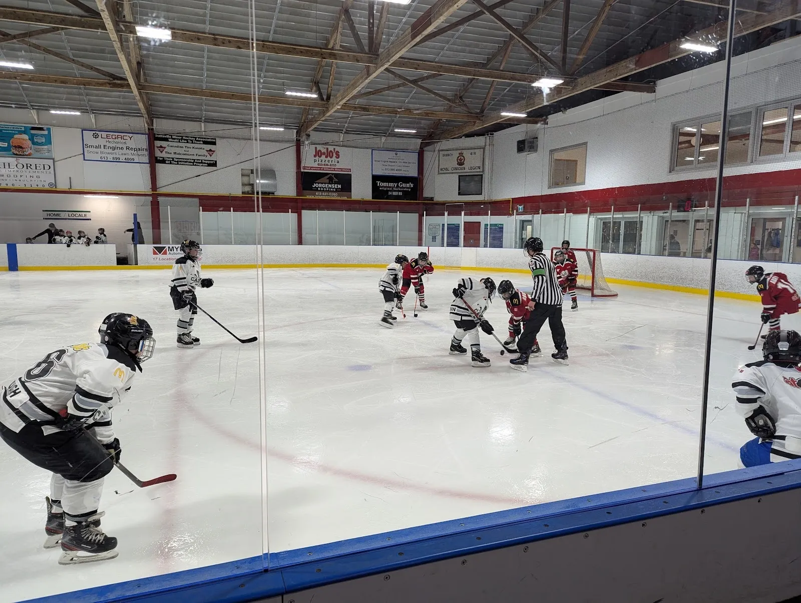 A faceoff at centre ice seen through the glass, players gathered around the circle beneath a steel-framed roof with red-trimmed far boards. — Johnny Leroux Stittsville Community Arena