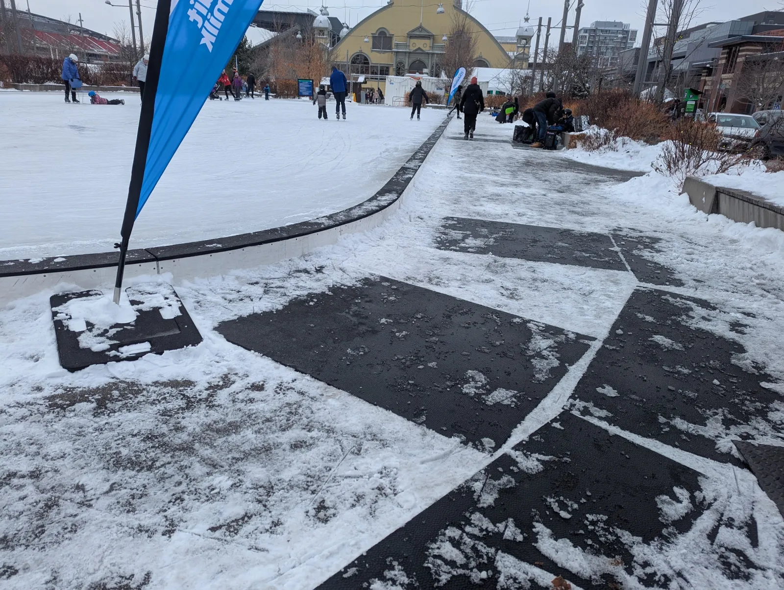 El circuito de patinaje de Lansdowne en una tarde de invierno, con una bandera azul en la orilla, patinadores en el hielo y el Aberdeen Pavilion detrás. — Lansdowne Park Outdoor Rink