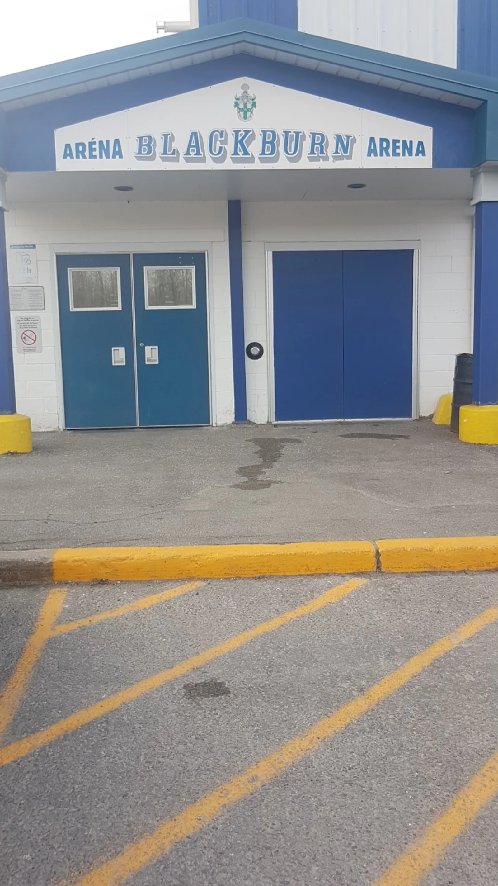 Entrada principal del edificio, con un letrero que dice "ARÉNA BLACKBURN ARENA" sobre puertas dobles azules y una puerta tipo cochera, frente a un estacionamiento. — Lois Kemp Arena (Blackburn)