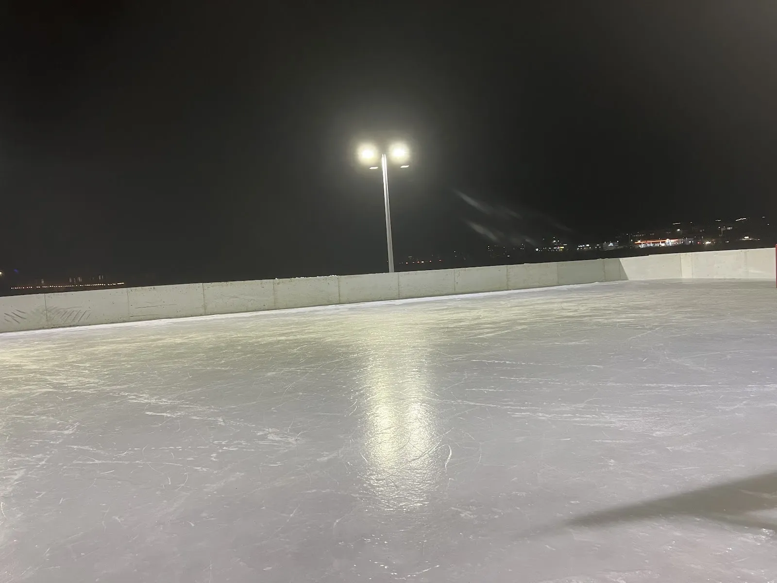 The Lyndon Hooper park rink at night, a wide empty sheet of ice inside white boards under floodlights with the lights of houses beyond. — Lyndon Hooper Park Outdoor Rink