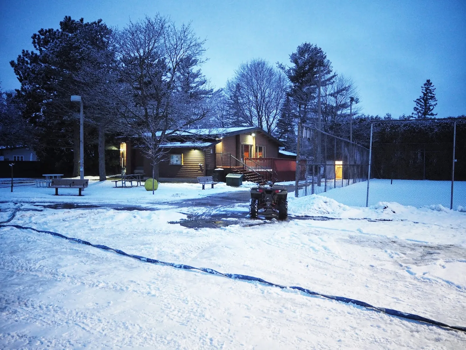 The Lynwood Village community building at dusk after a snowfall, lit windows behind the snowy pad and fenced courts. — Lynwood Park Outdoor Rink