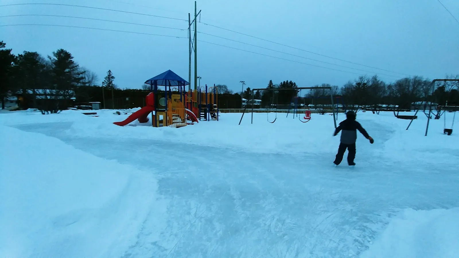 A lone skater on the Lynwood Park rink at dusk, the ice banked with snow and the playground behind. — Lynwood Park Outdoor Rink