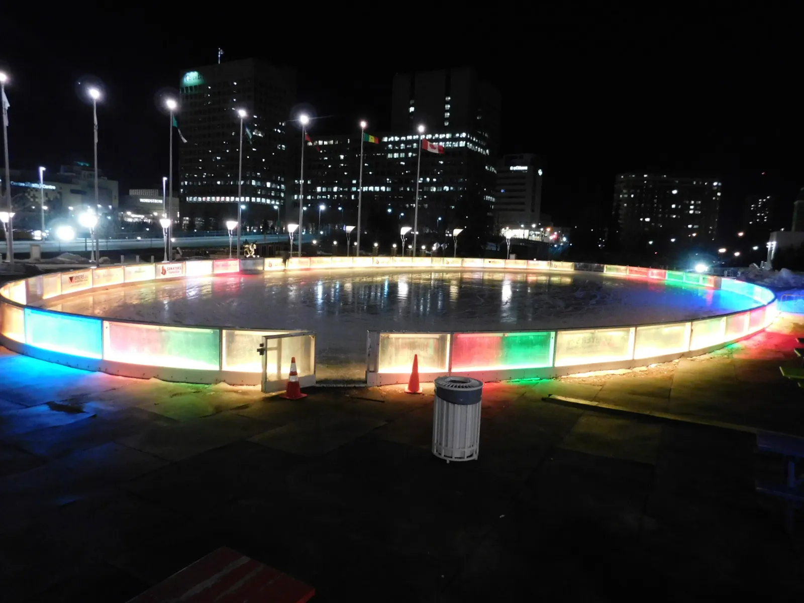 The oval Rink of Dreams ice sheet at night, ringed by blue, red and green lit boards, with downtown Ottawa office towers behind. — Marion Dewar Plaza (Rink of Dreams) Outdoor Rink