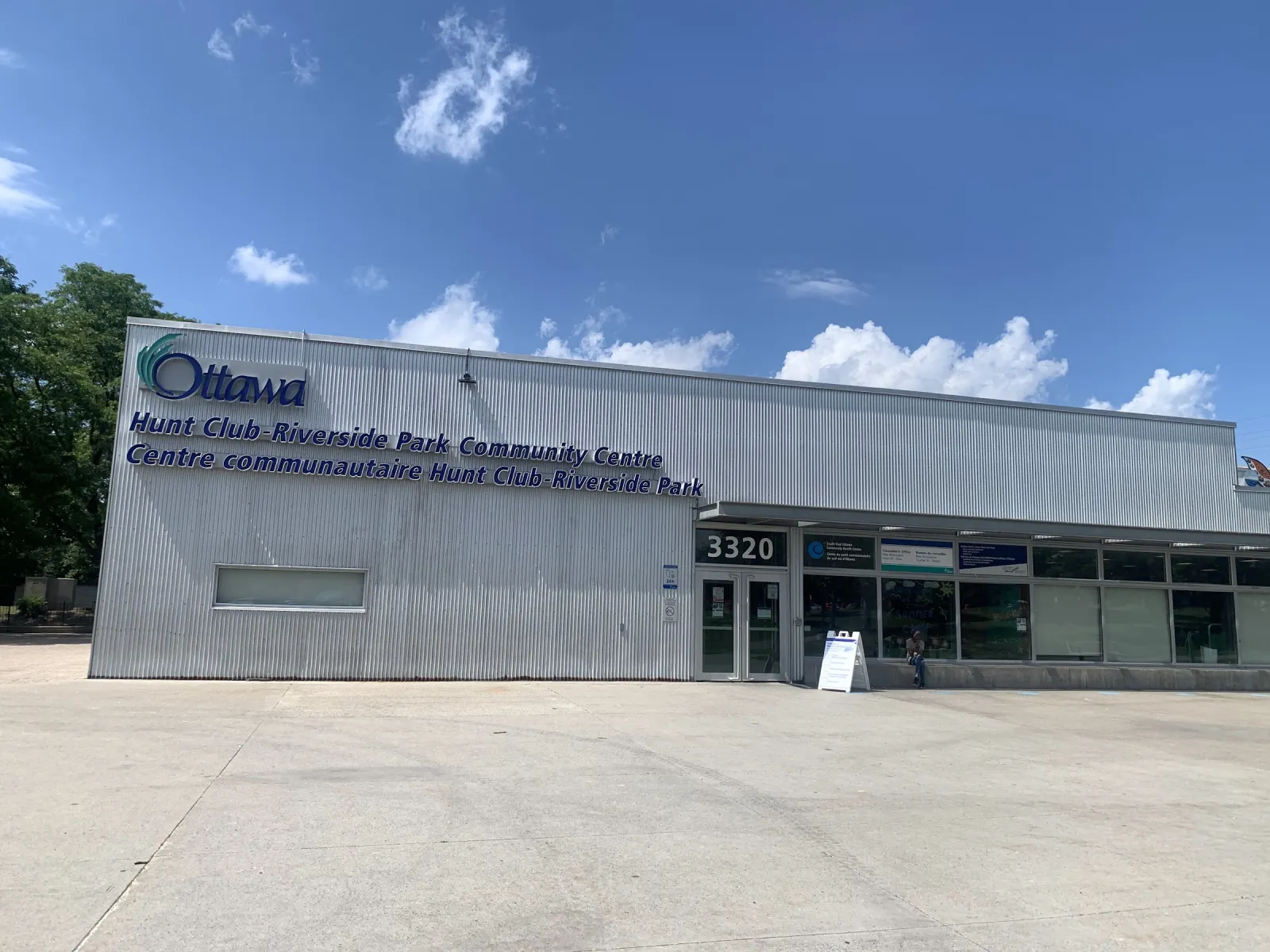 El Hunt Club Riverside Park Community Centre en 3320 Paul Anka Drive, un edificio bajo revestido de metal con entrada de vidrio, bajo un cielo azul. — McCarthy Park Outdoor Rink